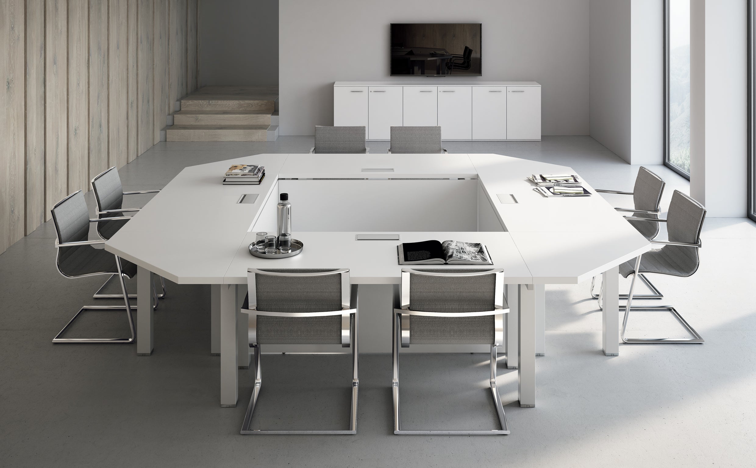 Une salle de conférence moderne est équipée d'une grande table de réunion carrée blanche Asset by Attitudes Bureaux, offrant la modularité, de huit chaises grises, de documents, d'une bouteille en verre avec des verres. Télévision, armoires, escalier et décor neutre complètent cet espace de travail modulable.