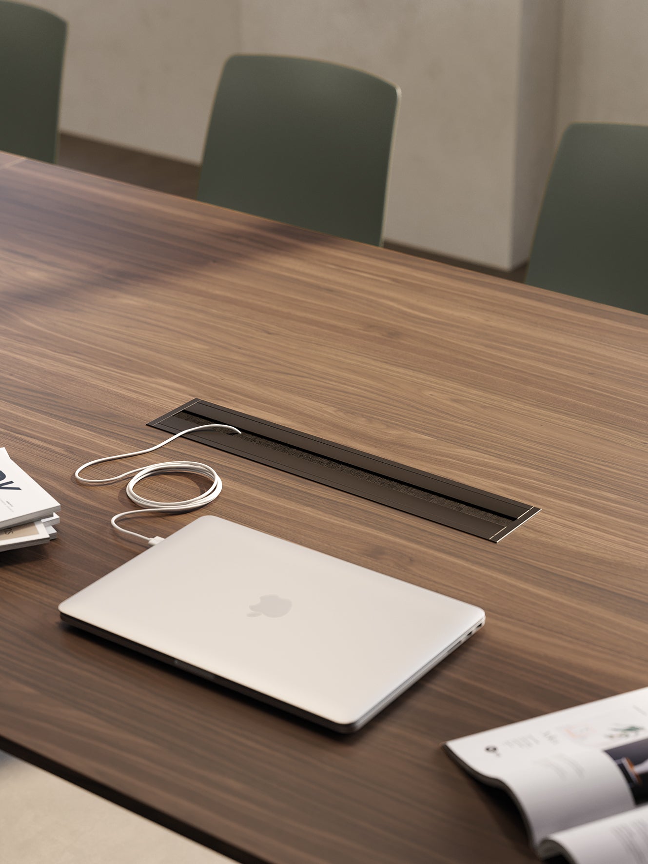 Un ordinateur portable argenté fermé avec un câble de chargement est posé sur une Table de réunion modulaires Be.1 de Progetto CMR, placée sur une table de conférence en bois à côté de papiers et de magazines, avec des chaises vertes visibles à l'arrière-plan.