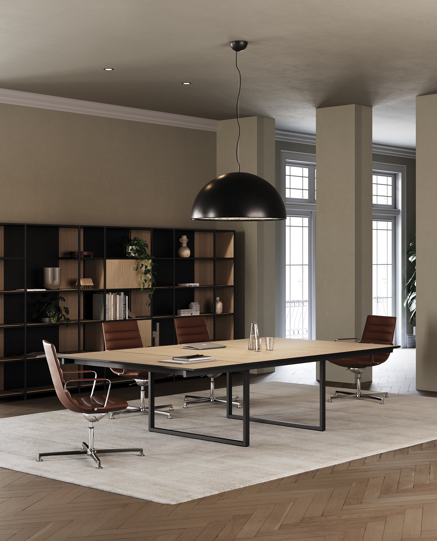 Une salle de conférence moderne avec une table rectangulaire modulaire en bois Be.1 de Progetto CMR, trois chaises pivotantes en cuir marron et des étagères avec des livres et de la décoration. De grandes fenêtres apportent de la lumière naturelle pour des réunions modulables.