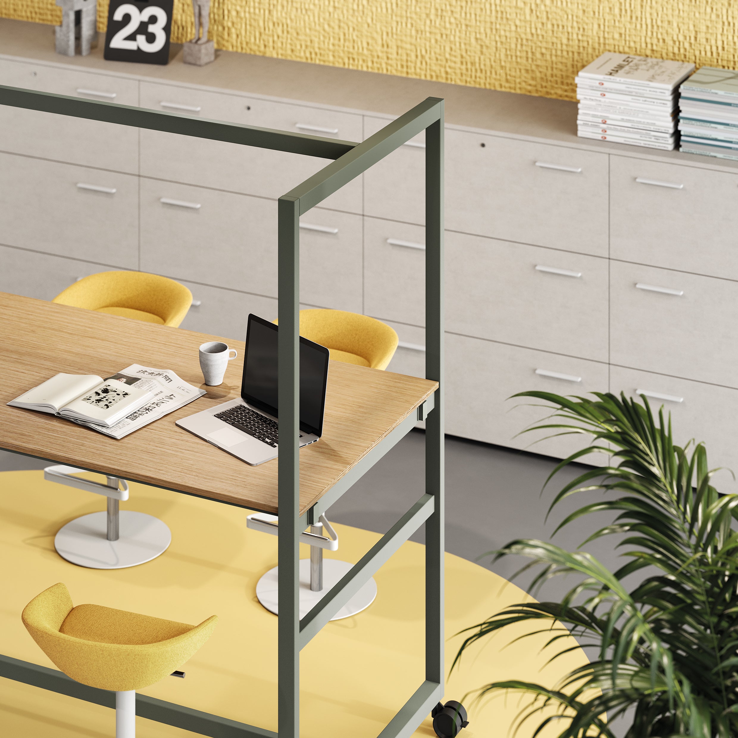 Espace de travail moderne comprenant la Table haute Nomad de Progetto CMR, deux chaises jaunes, un ordinateur portable, une tasse de café et des magazines. Des armoires blanches avec des tiroirs et des papiers à l'arrière-plan ; une plante au premier plan.