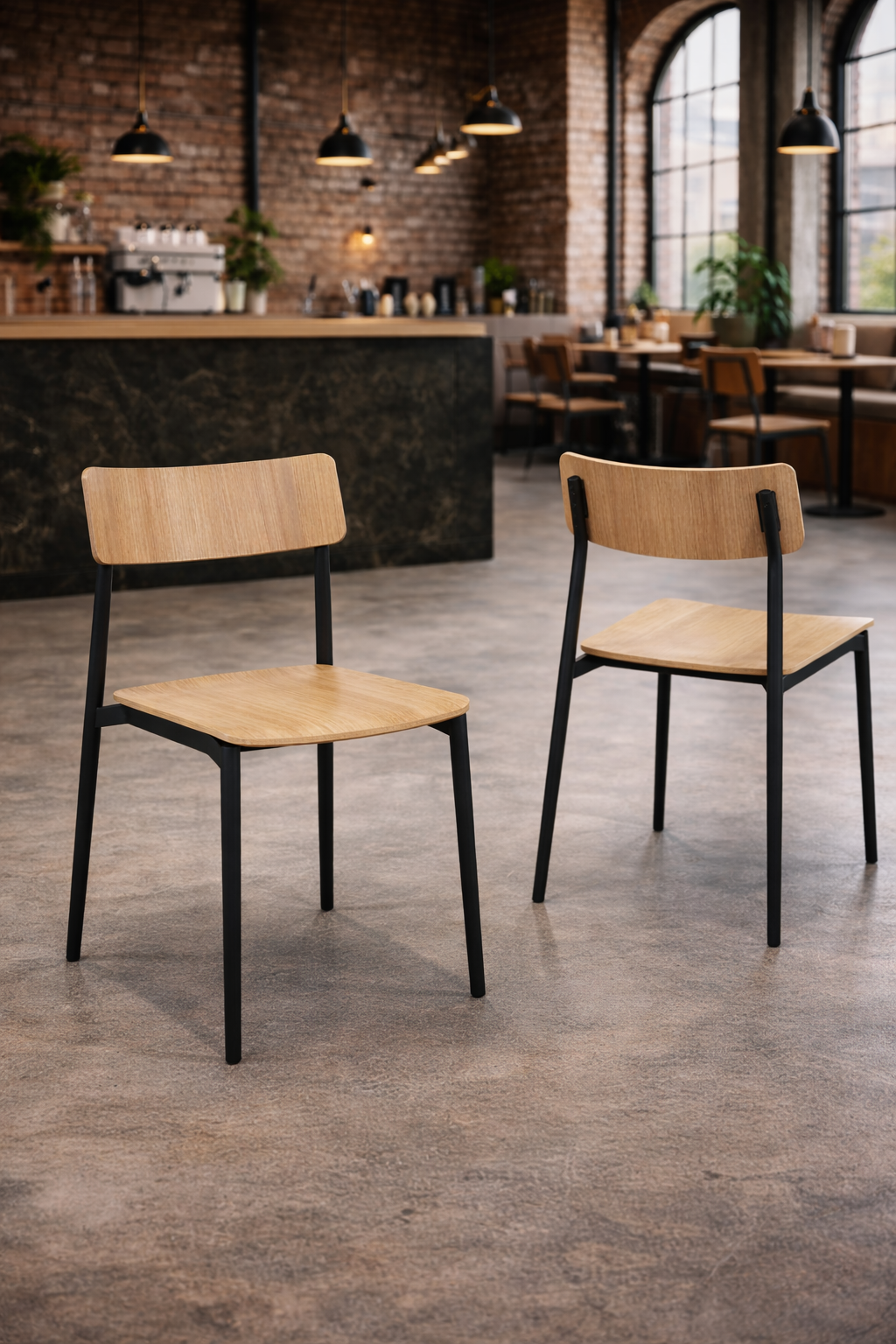 Deux chaises empilables Bois Cult d'Attitudes Bureaux, dotées d'une armature en métal noir et d'une assise en chêne courbé, sont posées sur un sol en béton dans un café moderne avec des murs en briques apparentes, des lampes suspendues, de grandes fenêtres cintrées et un comptoir à café.