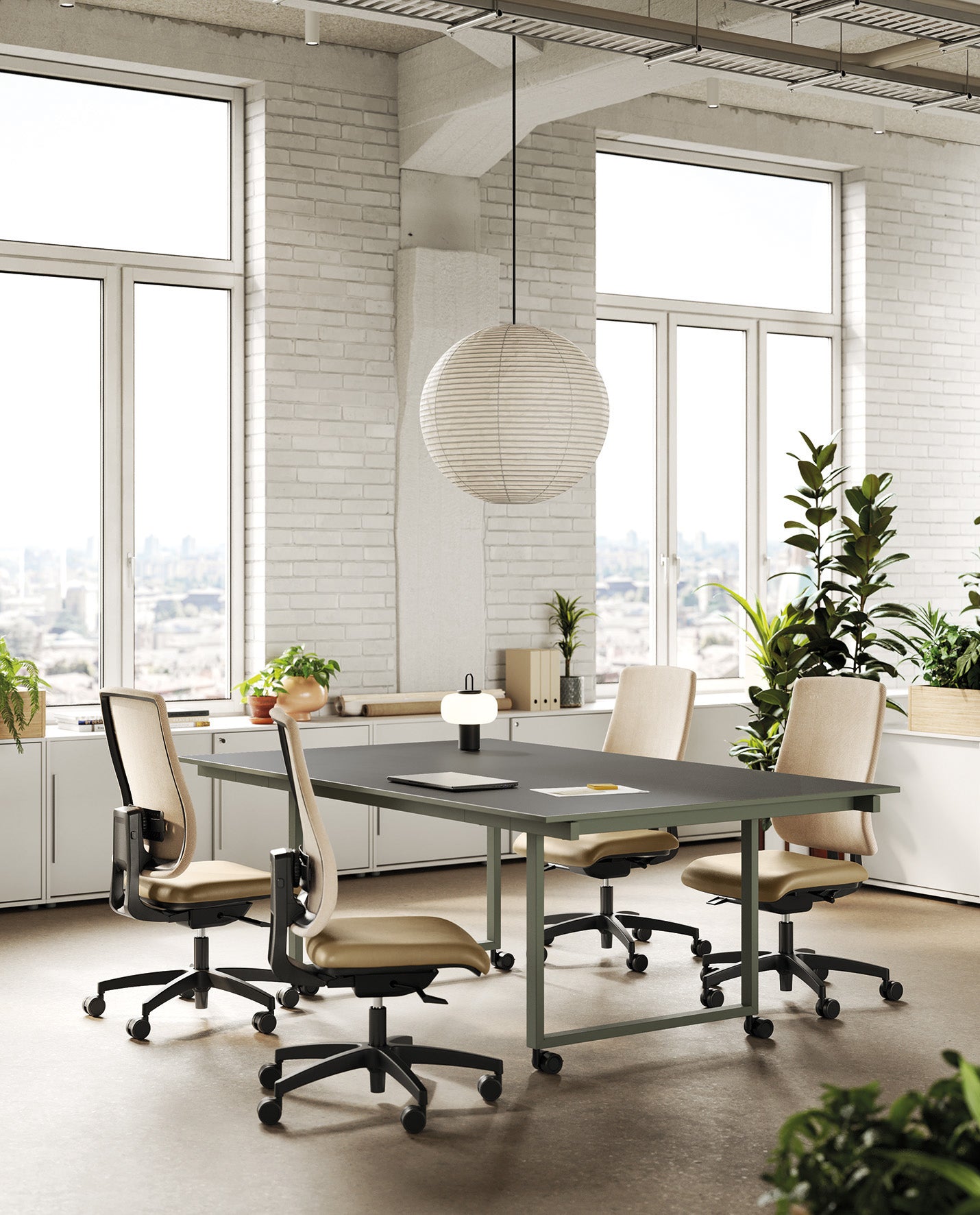 Un espace de bureau moderne avec quatre Chaises de bureau Denzel par Attitudes Bureaux autour d'une table sombre, de grandes fenêtres laissant entrer la lumière naturelle, des murs en briques blanches, des plantes d'intérieur et une lampe suspendue sphérique. Paysage urbain visible à l'extérieur.