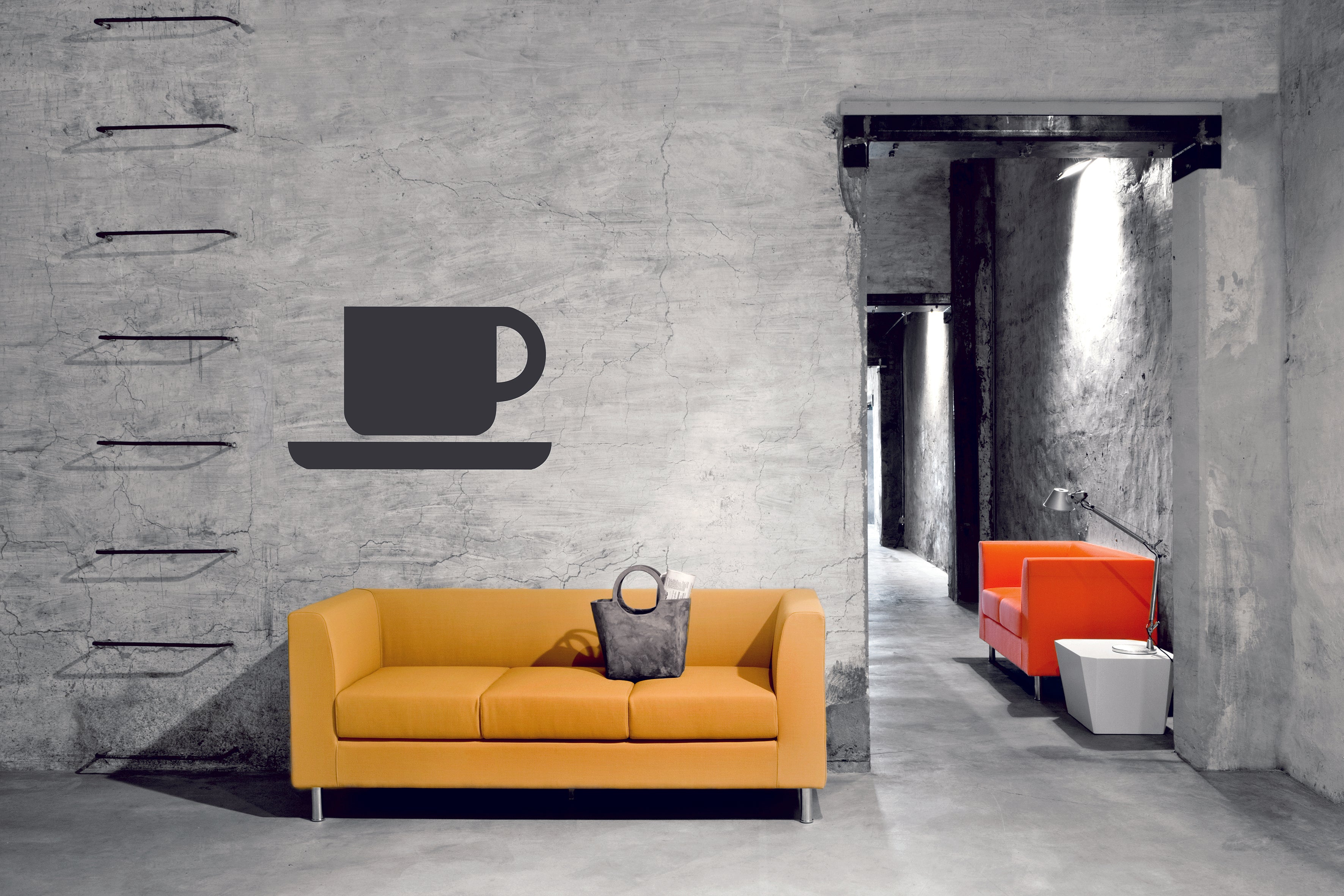 Une pièce industrielle moderne avec un domino Canapés jaune d'Attitudes Bureaux, un sac dessus, et un grand graphique de tasse et de soucoupe sur le mur en béton ; à côté, des espaces lounge avec une chaise orange et une table d'appoint blanche sur des murs gris.