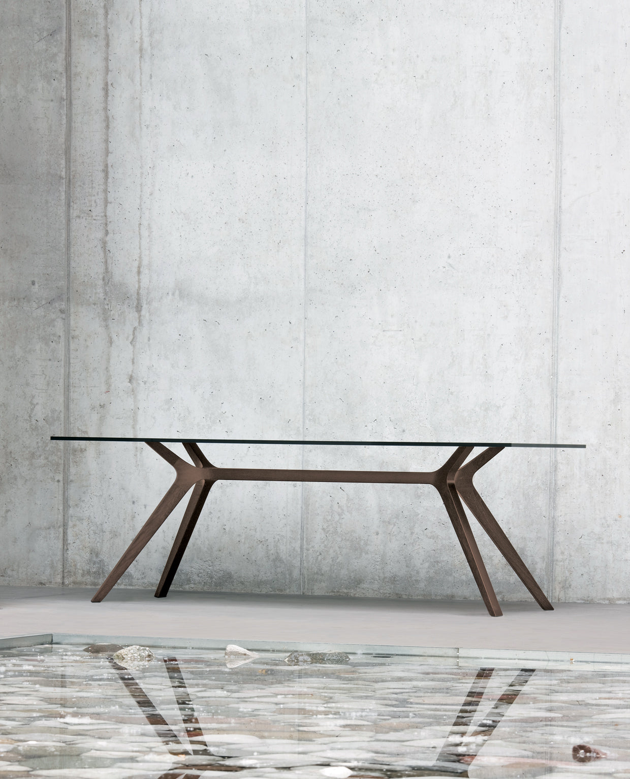 Une table de réunion Dr moderne de Claudio Bellini, avec un plateau en verre et des pieds en bois inclinés, se dresse devant un mur en béton gris clair, dont l'élégante surface laisse apparaître des coquillages et des pierres.