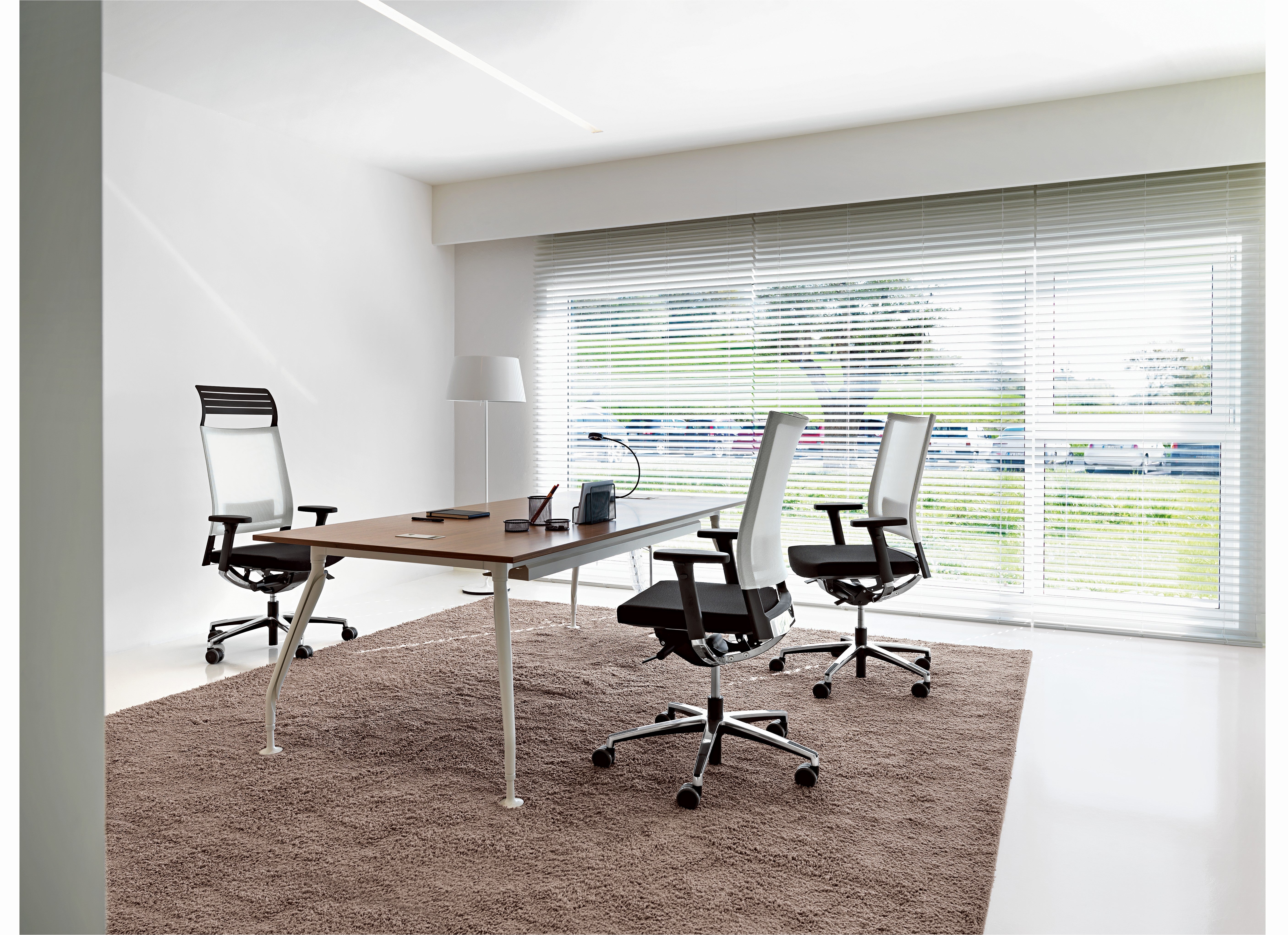Bureau moderne avec un grand bureau en bois, trois chaises Fauteuil ergonomique Graphic de Baldanzi & Novelli Design, une lampe et un ordinateur portable sur un tapis marron. Les fenêtres du sol au plafond avec stores laissent entrer la lumière naturelle et révèlent la verdure à l'extérieur.