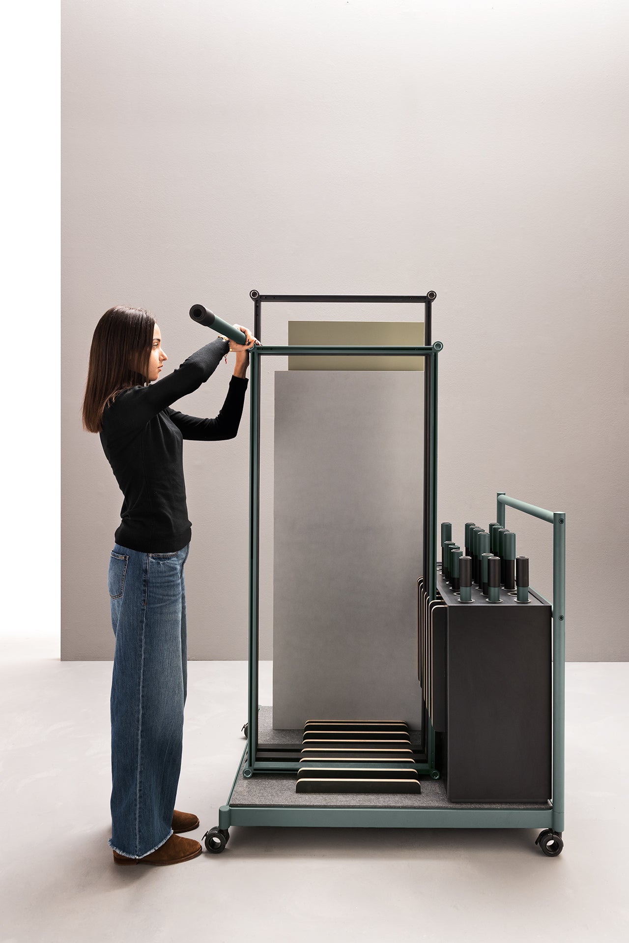 Une femme en jean et haut noir se tient à côté du Fluido, mobilier modulaire transformable d'Attitudes Bureaux, et retire un cylindre noir d'une fente. Le système reconfigurable organise efficacement les objets et les panneaux similaires.