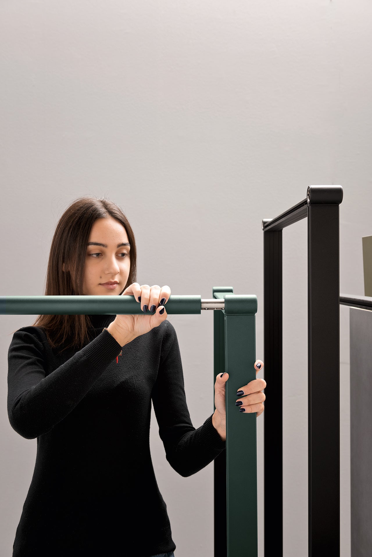 Une femme aux longs cheveux noirs et au haut noir assemble le Fluido mobilier modulaire transformable d'Attitudes Bureaux, en connectant des barres vertes et noires dans un montage sans outil sur un fond gris clair uni.