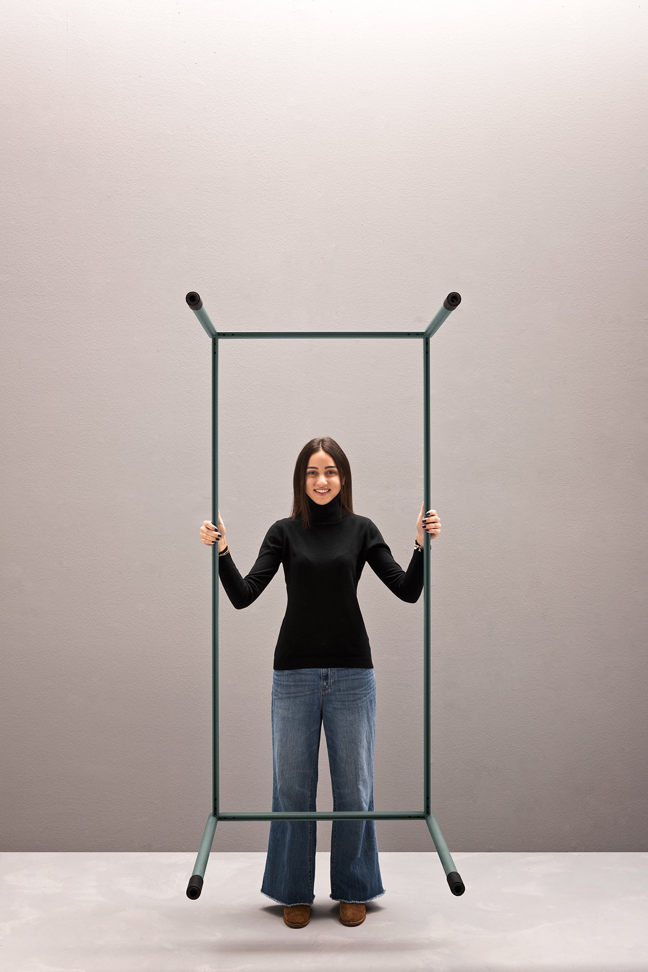 Une femme en col roulé noir et jean bleu sourit à l'intérieur d'un cadre métallique rectangulaire, présentant Fluido, mobilier modulaire transformable d'Attitudes Bureaux, facile à assembler sans outils, sur un fond gris uni.