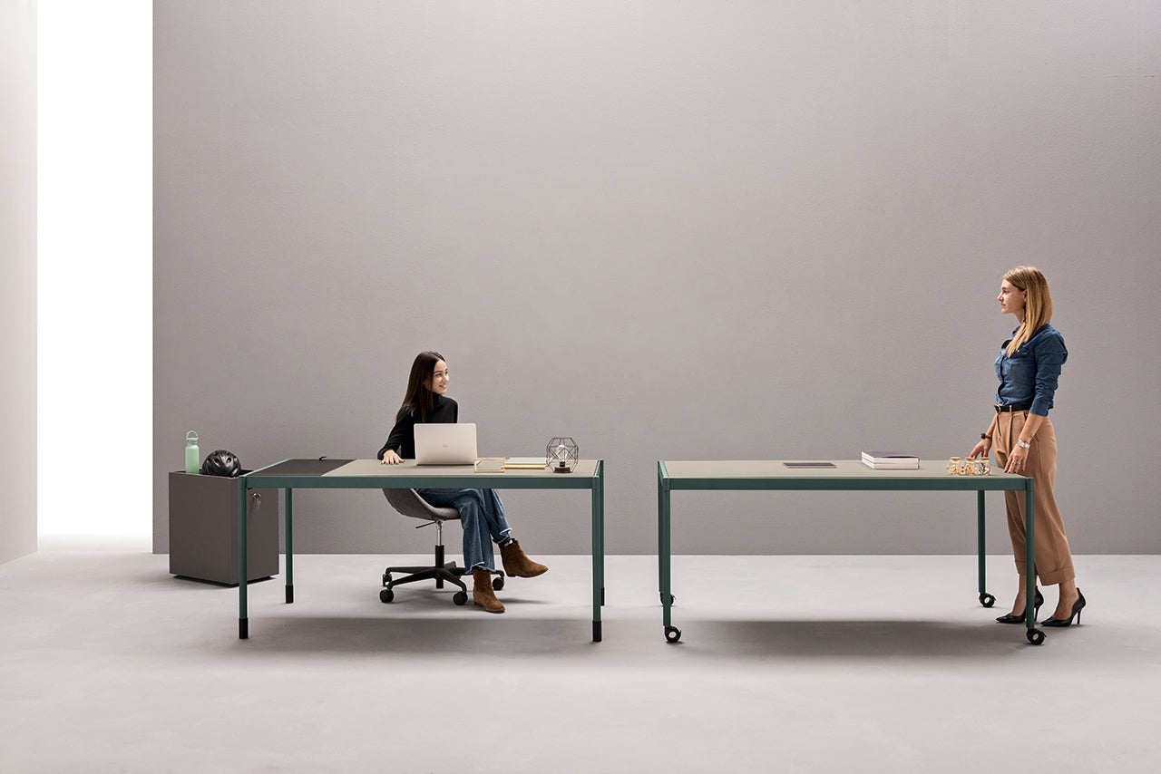 Dans un bureau minimaliste, deux femmes utilisent Fluido, mobilier modulaire transformable d'Attitudes Bureaux, l'une à un bureau avec un ordinateur portable, l'autre debout à proximité. L'espace lumineux est doté d'un mobilier modulaire simple dans des tons neutres.