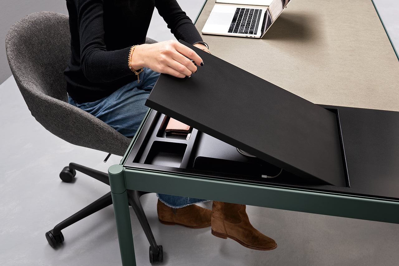 Une personne portant un jean, des bottes et un haut noir est assise à un Fluido, mobilier modulaire transformable par Attitudes Bureaux, ouvre un compartiment de bureau caché contenant de petits objets. Un ordinateur portable est posé à proximité sur le bureau.