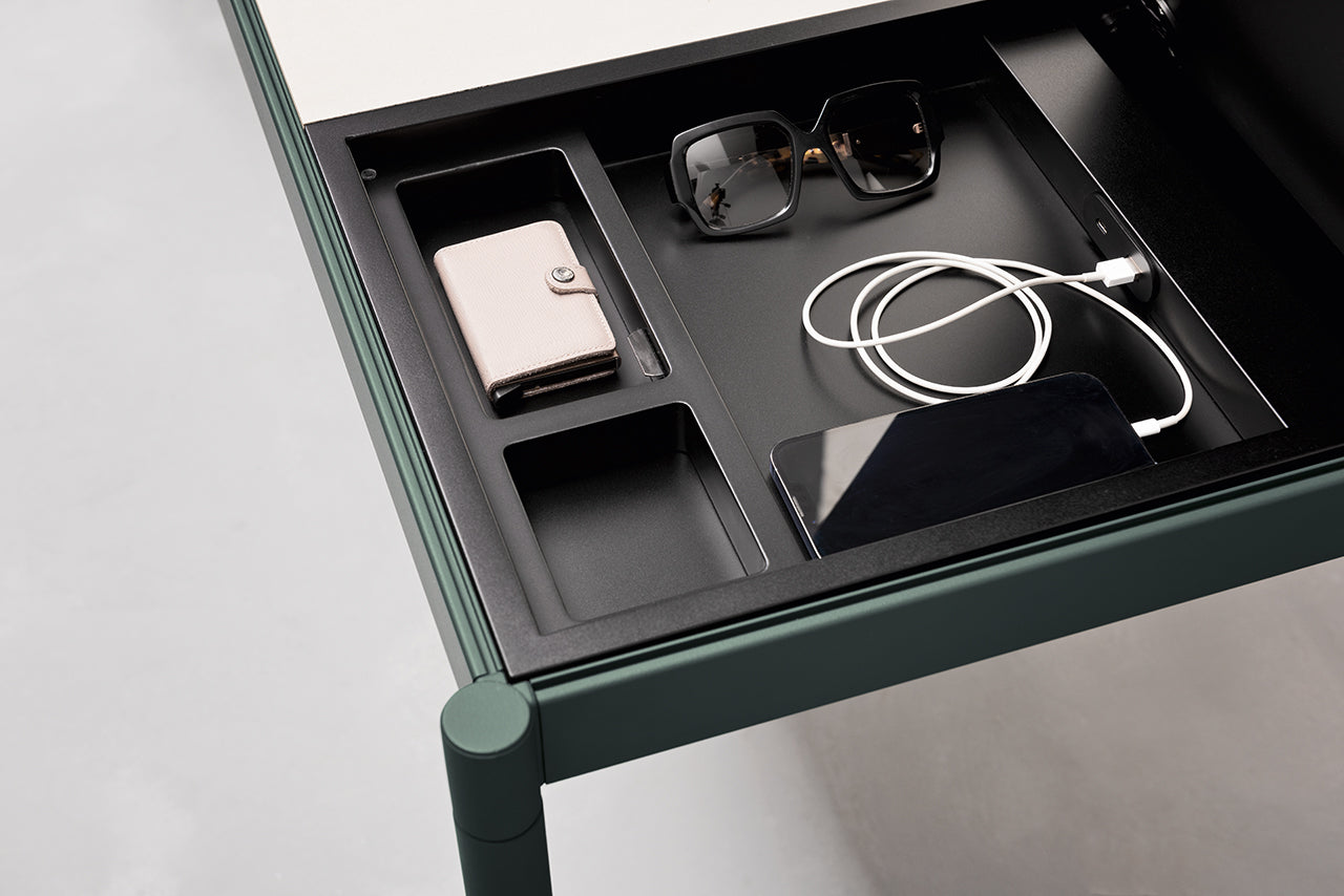 Gros plan sur le Fluido mobilier modulaire transformable d'Attitudes Bureaux : un portefeuille rose, des lunettes de soleil noires et un smartphone avec un câble de recharge dans des compartiments séparés. Le bureau à l'armature verte est doté d'un plateau de couleur claire.