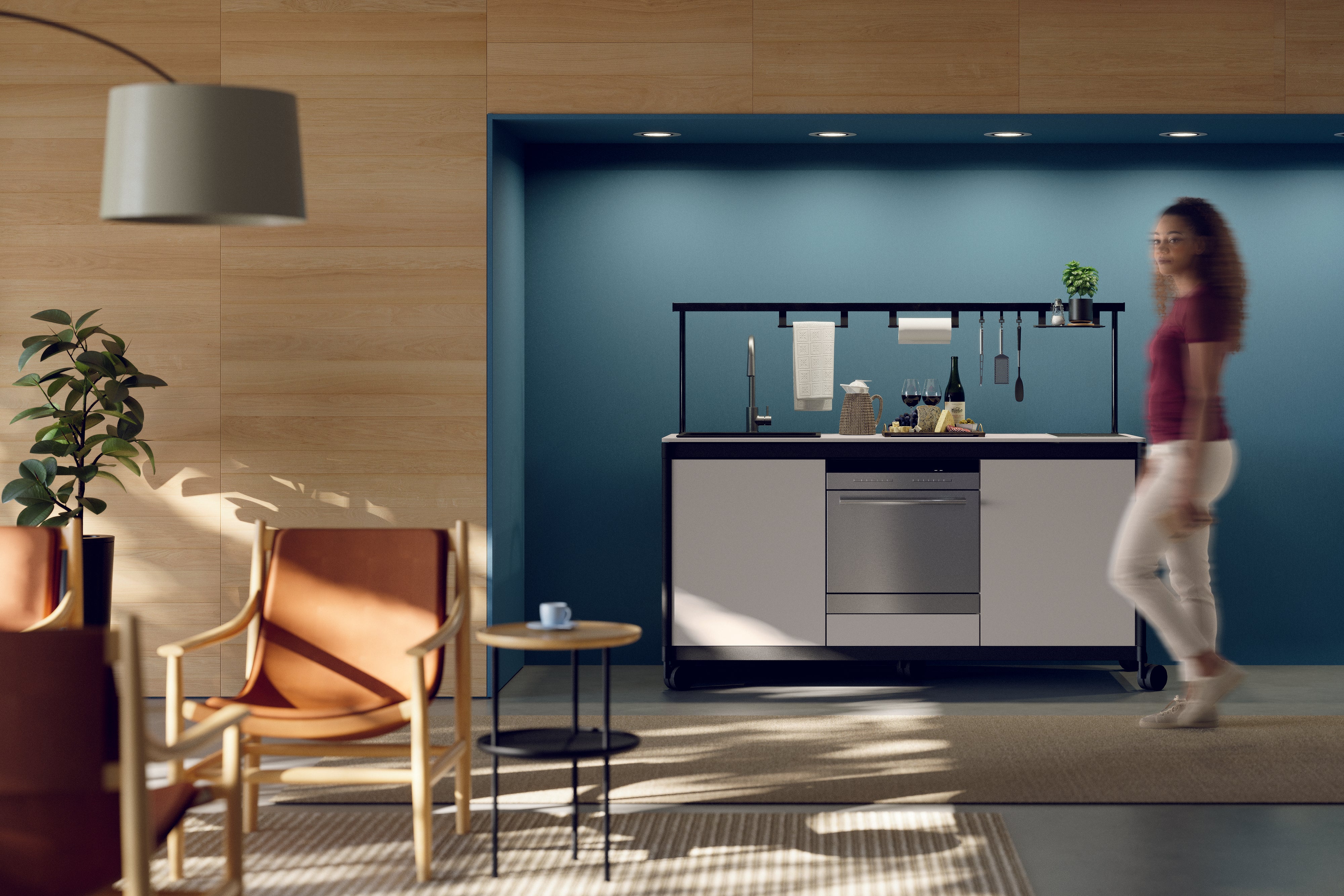 Une femme passe devant une kitchenette Fluido® d'Attitudes Bureaux, composée d'armoires blanches, d'un évier et d'ustensiles sur un mur bleu. Des murs lambrissés, des plafonniers et des chaises de bureau en cuir avec une table d'appoint complètent l'espace pause moderne.