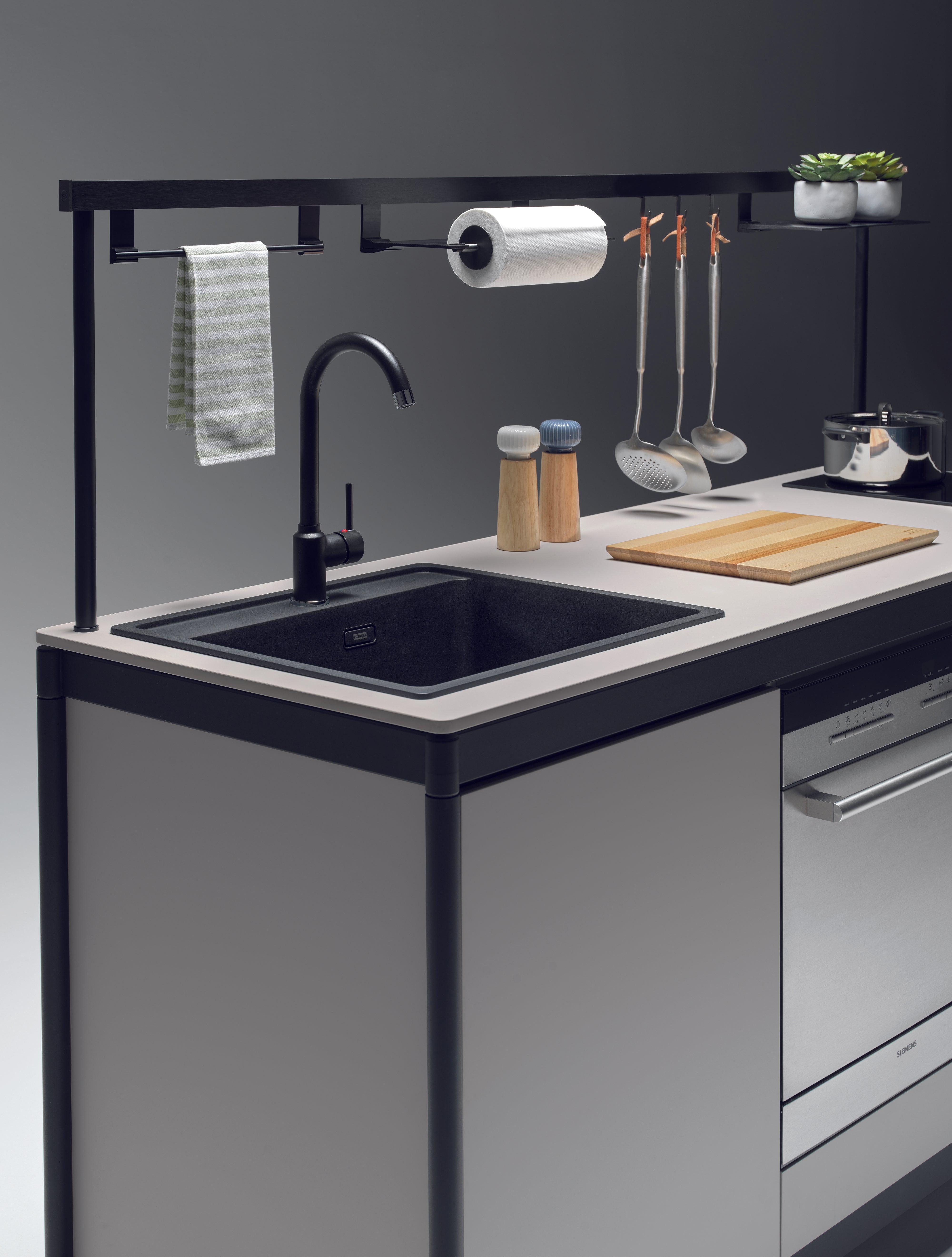 La kitchenette Fluido® d'Attitudes Bureaux est composée d'un évier épuré avec un robinet noir, d'ustensiles suspendus, d'une serviette, d'un rouleau d'essuie-tout, de moulins à sel et à poivre, d'une planche à découper, d'une casserole et d'une petite plante sur le plan de travail.