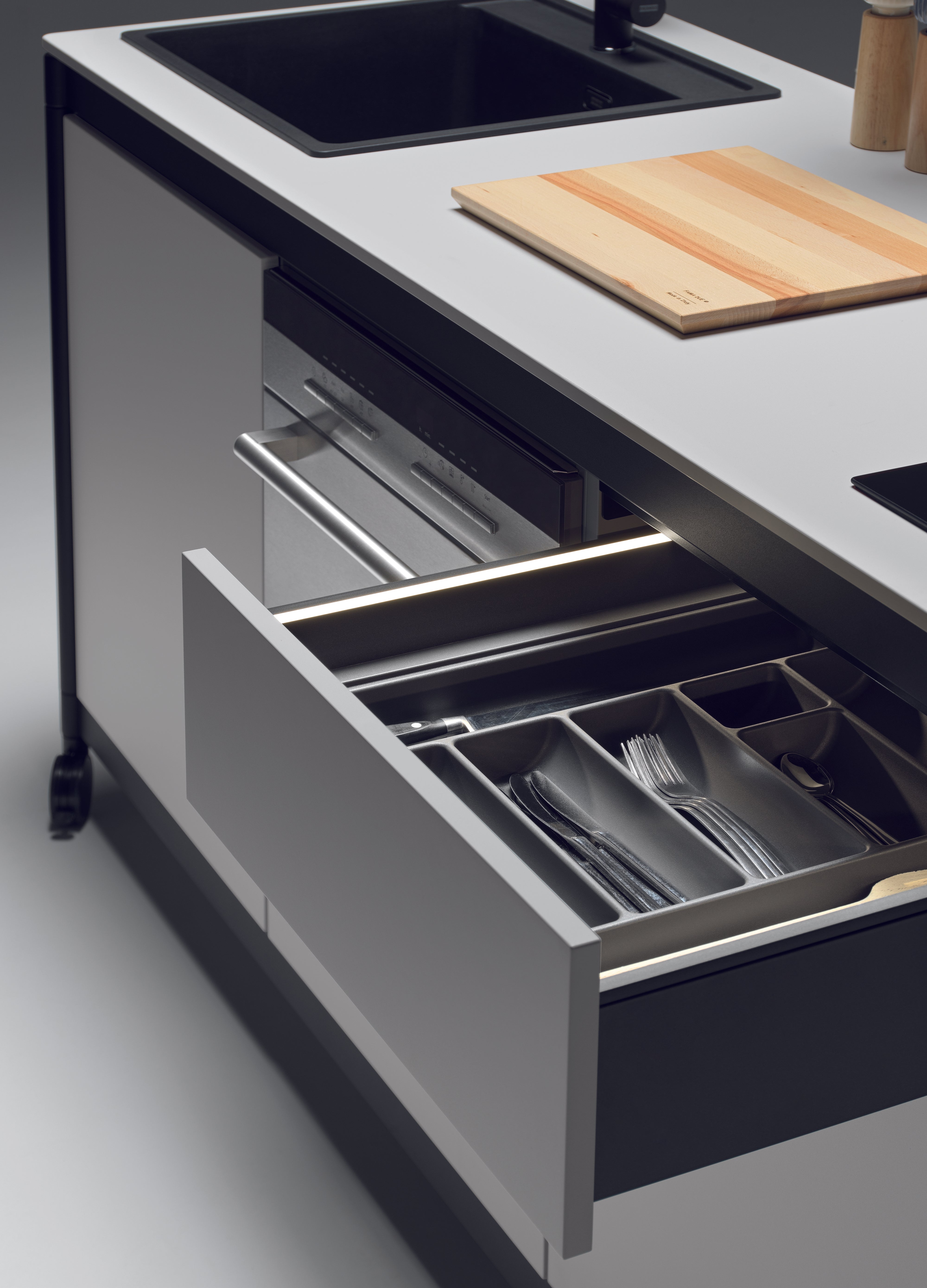 La kitchenette Fluido® d'Attitudes Bureaux comprend un îlot moderne avec un tiroir ouvert pour ranger les couverts, un évier, une planche à découper en bois sur le plan de travail et un meuble minimaliste aux lignes épurées - idéal pour toute pause cuisine bureau ou kitchenette espace.