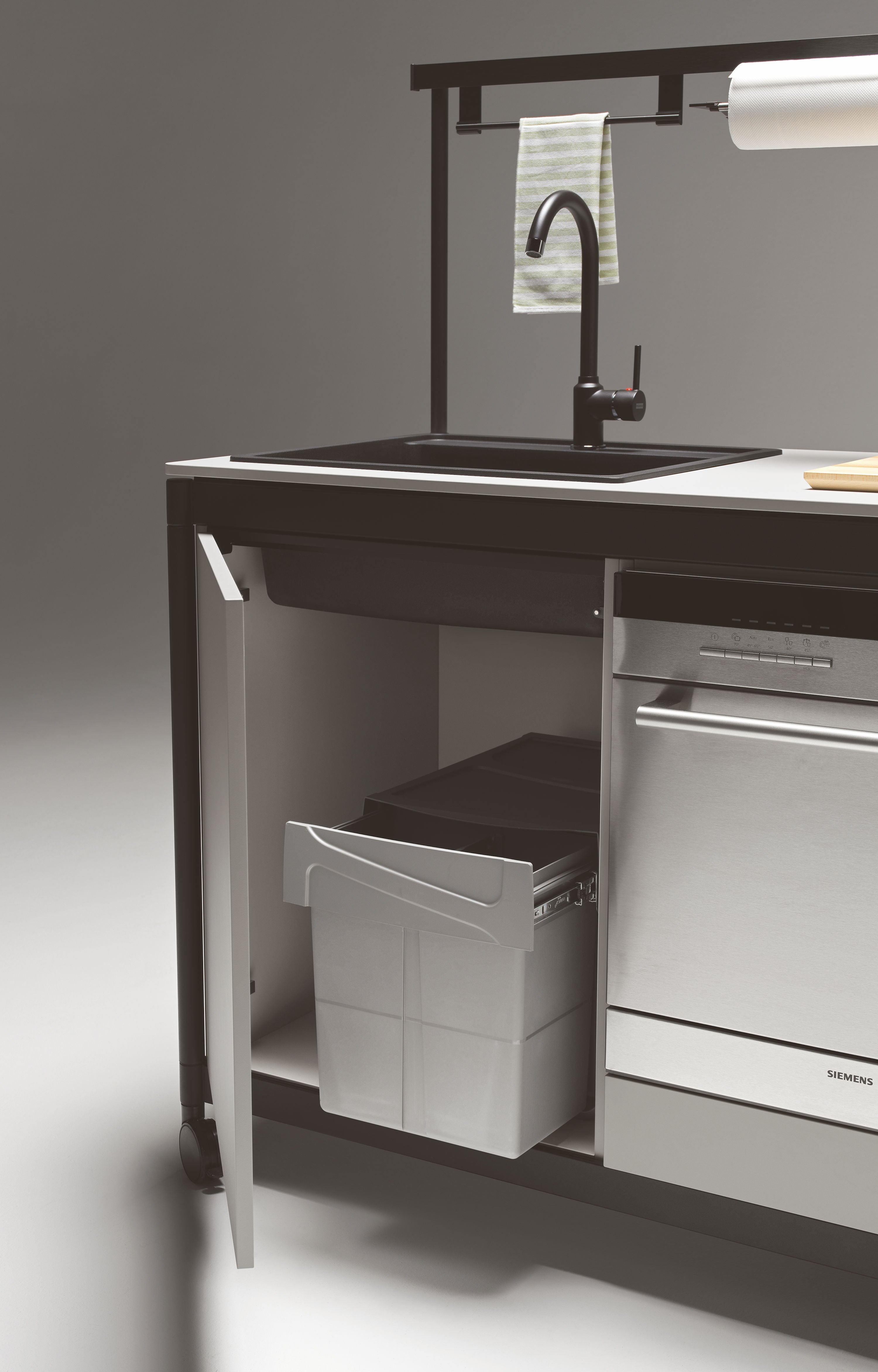 La kitchenette Fluido® d'Attitudes Bureaux est équipée d'un évier moderne à robinet noir, d'une serviette rayée au-dessus et d'un meuble ouvert au-dessous avec une poubelle extractible à côté d'un lave-vaisselle en acier inoxydable.