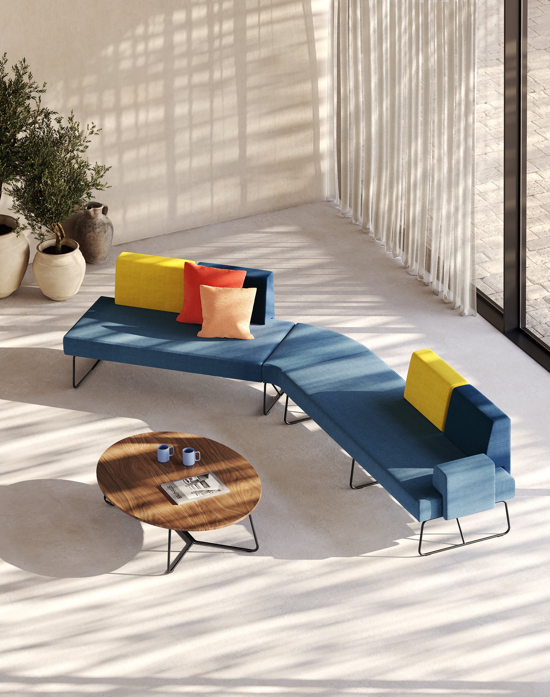 Un salon moderne avec le canapé sectionnel Fedra bleu de d.FLUX Canapés d'accueil, des coussins d'appoint jaunes et orange, une table basse ronde en bois avec des tasses et un magazine, des plantes en pot et la lumière du soleil à travers des rideaux diaphanes.