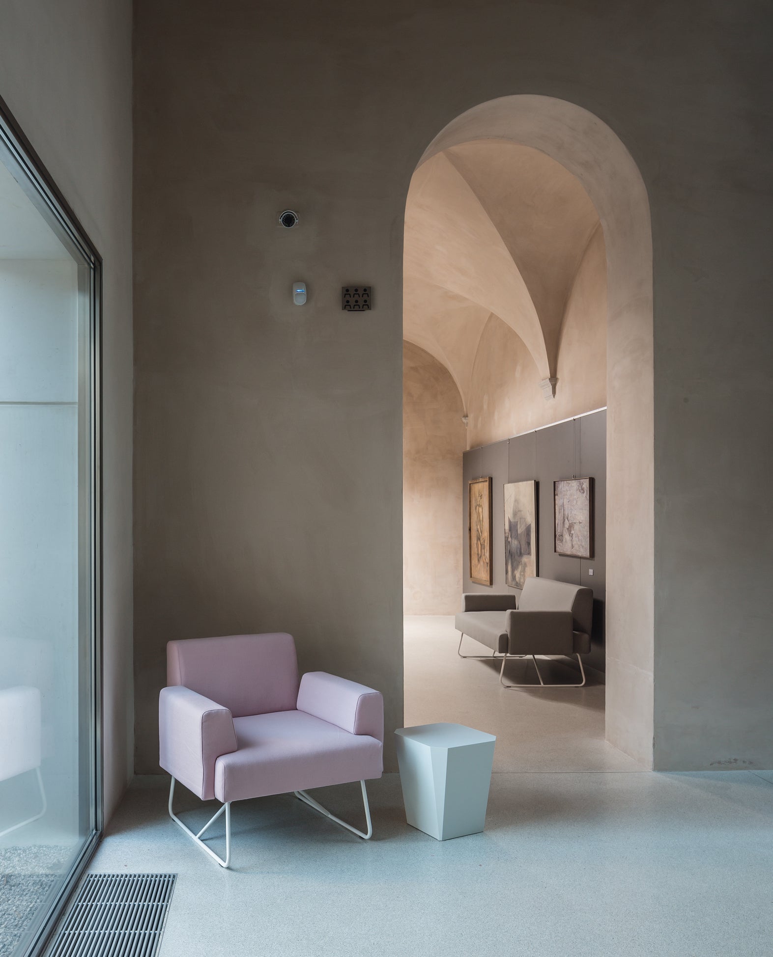 Un fauteuil Canapés d'accueil Fedra de d.FLUX en rose pastel et une petite table blanche sont placés près d'une paroi en verre dans une pièce minimaliste aux plafonds voûtés. Au-delà d'une arche, on peut voir d'autres sièges et des œuvres d'art encadrées.