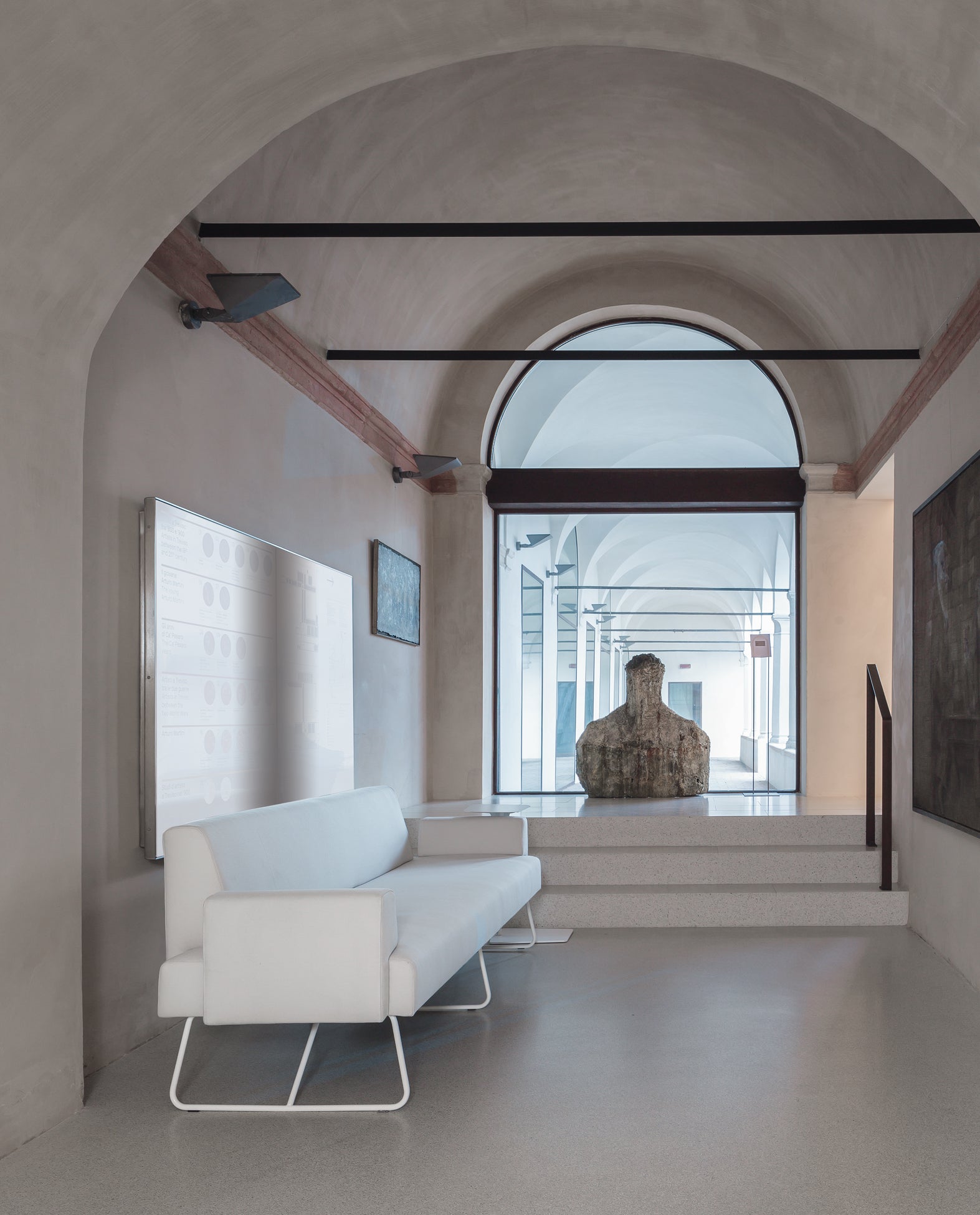 Un couloir moderne avec des plafonds voûtés, des murs clairs, un banc d.FLUX Canapés d'accueil Fedra blanc, un grand présentoir mural et une sculpture au bout, près d'une porte vitrée où la lumière naturelle pénètre à flots.