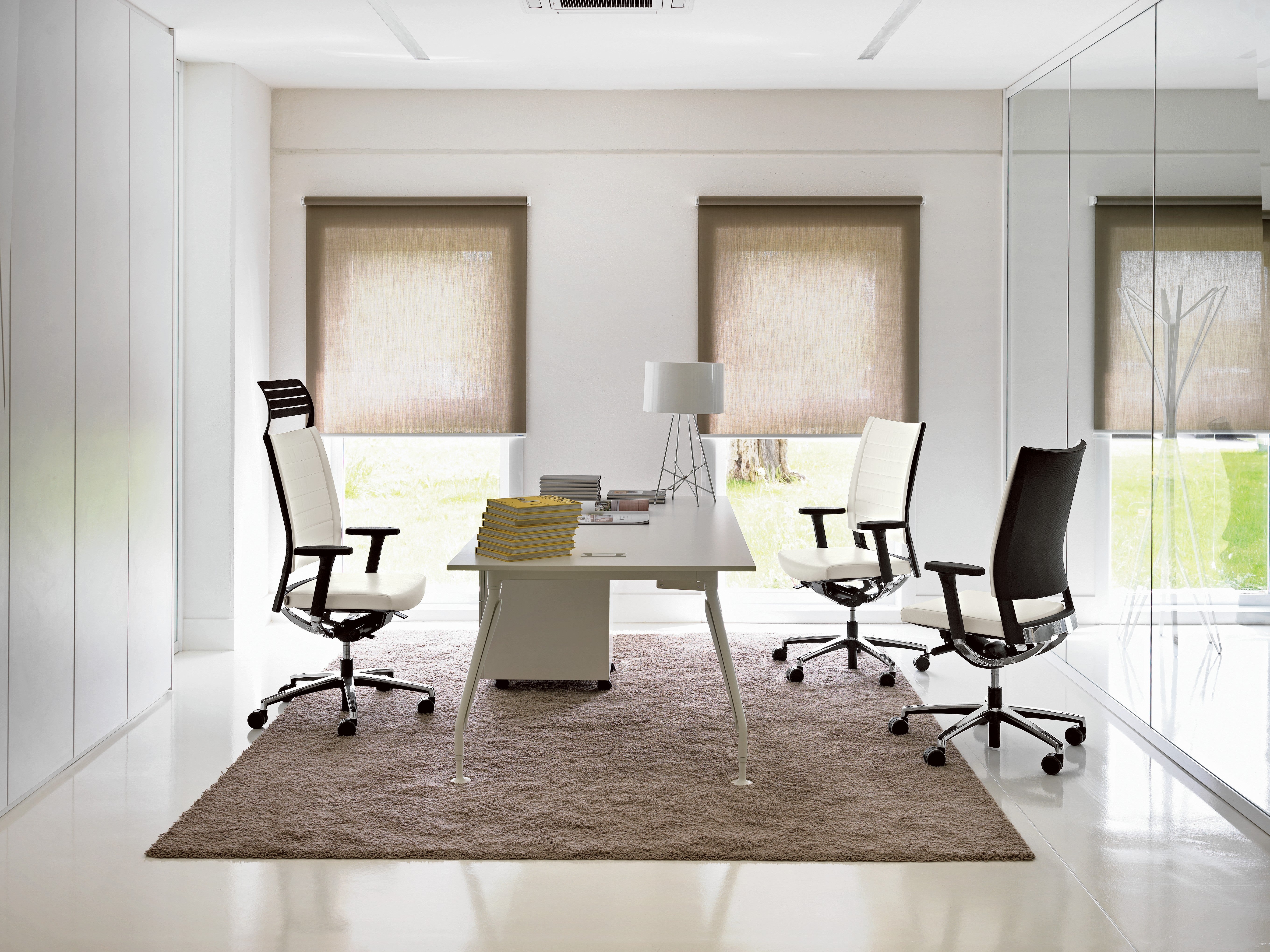Bureau moderne avec un bureau blanc, trois Fauteuils ergonomiques Graphic de Baldanzi & Novelli Design, une lampe, des livres et de la paperasse sur un tapis beige ; de grandes fenêtres avec des stores laissent entrer une lumière naturelle douce pour un look contemporain.