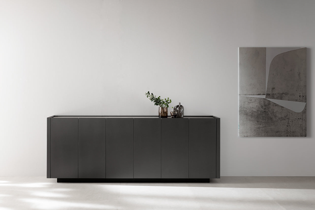 Un intérieur minimaliste présente le buffet Kyo noir mat d'Attitudes Bureaux contre un mur blanc. Un vase et un service à thé sont posés dessus, tandis qu'un tableau abstrait monochrome est accroché à droite. Fabriqué en matériaux haut de gamme.