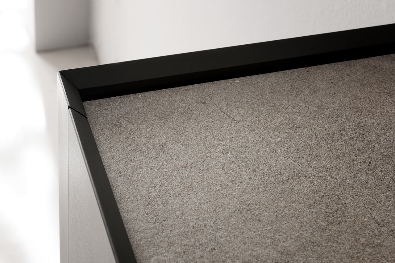 Gros plan sur l'angle supérieur de Kyo d'Attitudes Bureaux, doté d'un cadre métallique noir et d'une surface en pierre grise texturée - des matériaux de première qualité idéaux pour les bureaux de direction, présentés contre un mur blanc.