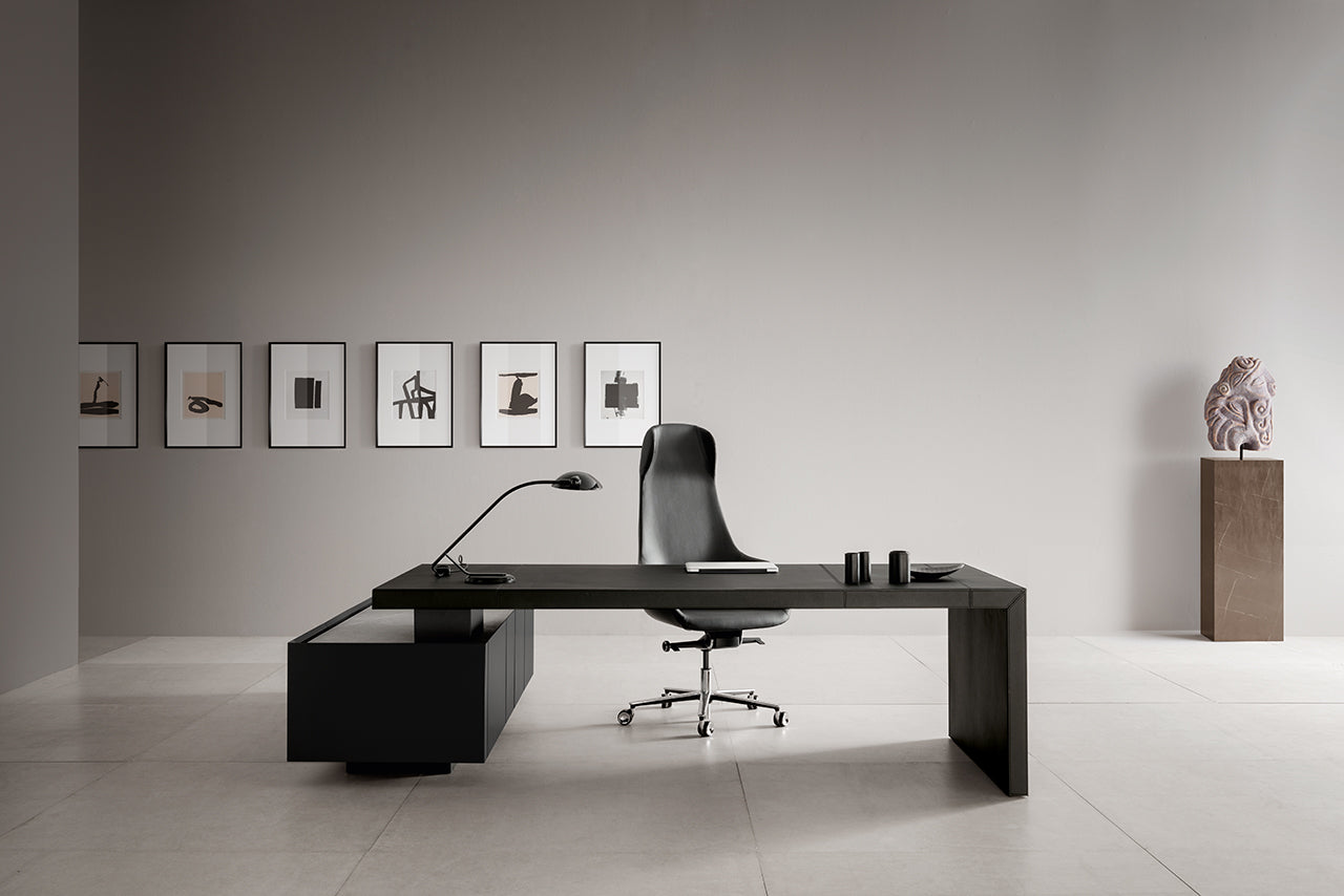Ce bureau minimaliste comprend le bureau Kyo d'Attitudes Bureaux, une chaise à dossier haut, une lampe de bureau et des fournitures de bureau. Des œuvres d'art sont accrochées au mur gris derrière, tandis qu'une sculpture est posée sur un piédestal dans le coin.