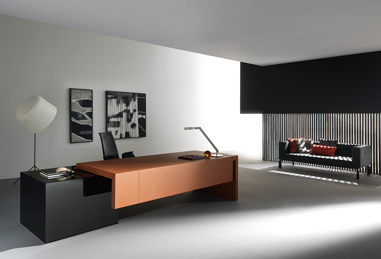 Un bureau moderne avec le bureau Kyo d'Attitudes Bureaux en marron et noir, une chaise noire, deux peintures murales abstraites, un lampadaire géométrique, ainsi qu'un canapé noir avec des coussins rouges et gris près des lattes verticales qui laissent passer la lumière.