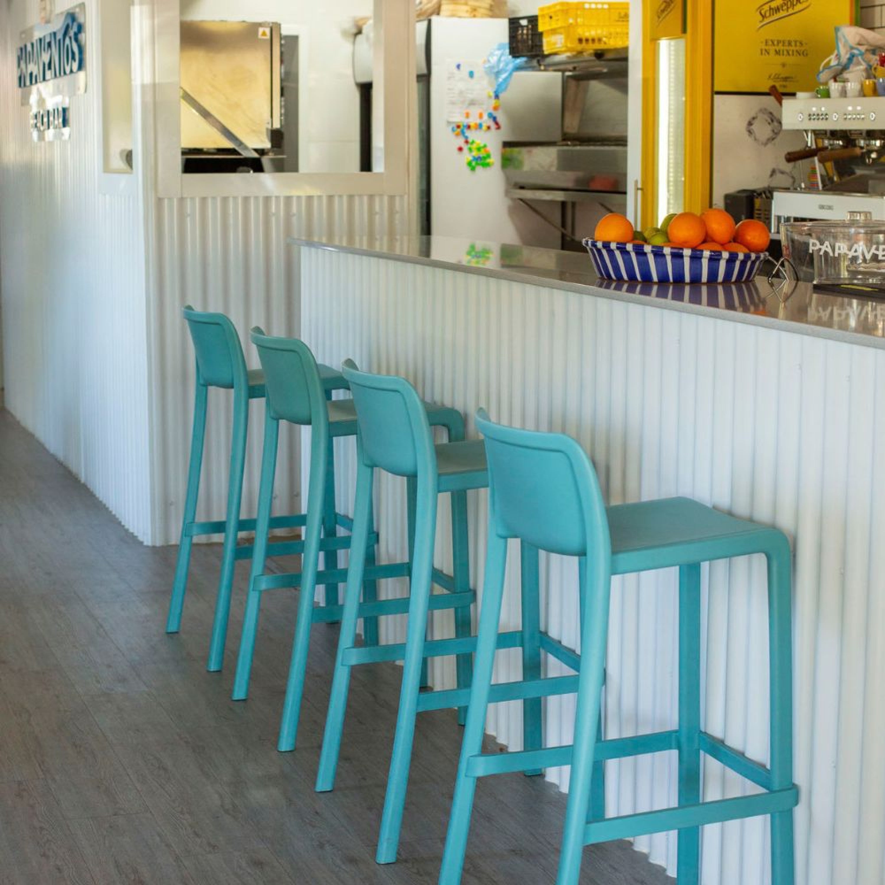 Un comptoir de café moderne présente quatre Tabourets ATTIC d'Attitudes Bureaux en turquoise, un bol d'oranges et une machine à café. Les murs d'un blanc éclatant et le sol en bois créent une atmosphère accueillante.
