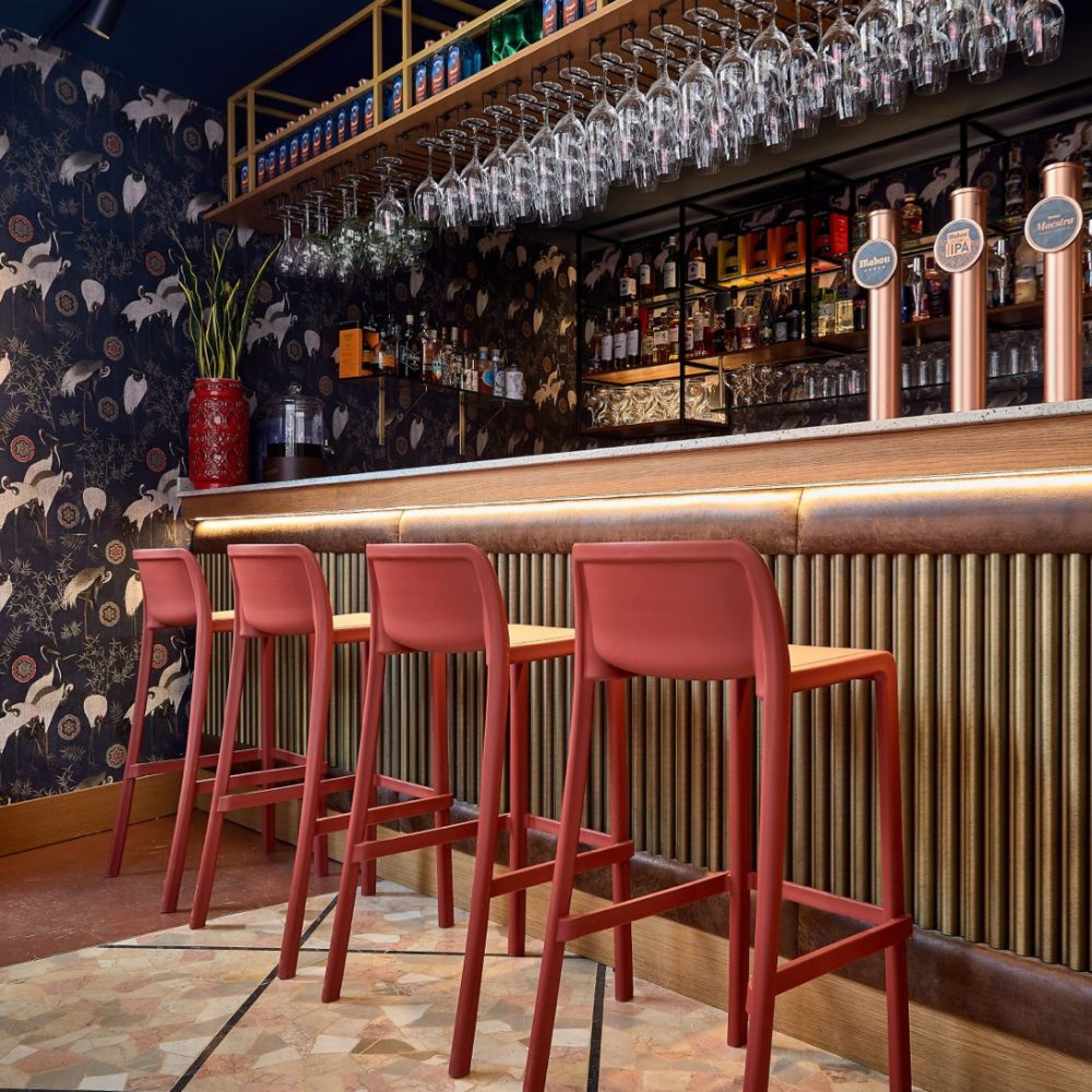 Un bar élégant présente quatre Tabourets ATTIC d'Attitudes Bureaux, en polypropylène rouge, sur un comptoir à la façade dorée nervurée. Des verres suspendus, des bouteilles d'alcool et des robinets de bière décorent l'espace aux côtés d'un papier peint à motifs et d'un grand vase rouge contenant des plantes.