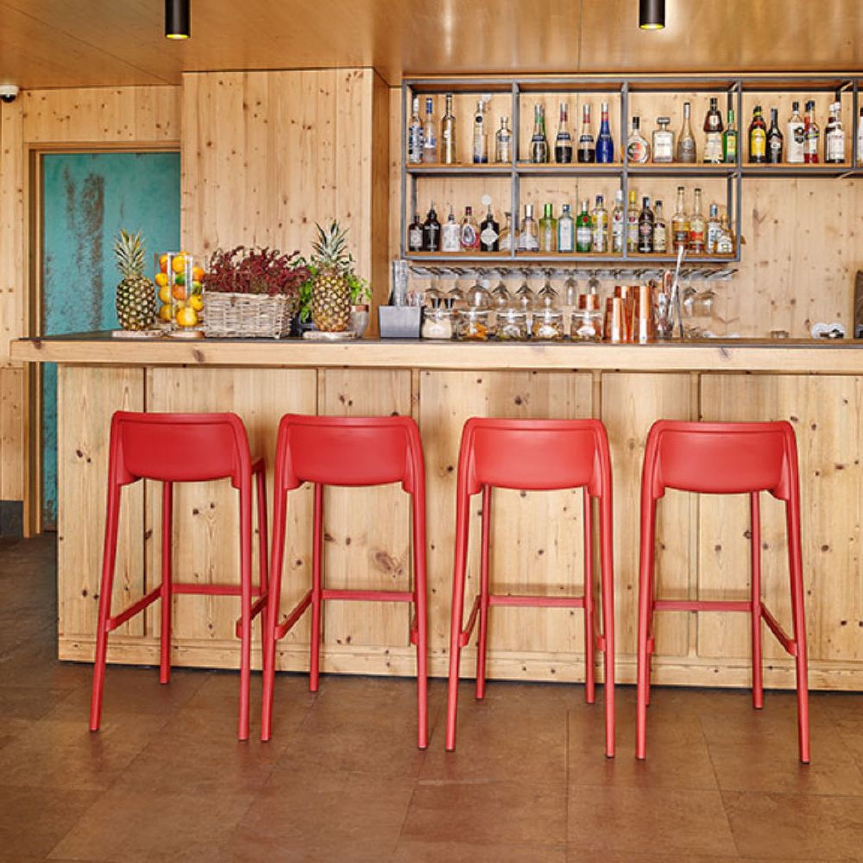 Un bar en bois avec quatre Tabourets ATTIC by Attitudes Bureaux rouge vif devant, des bouteilles et des verres assortis sur des étagères derrière, et un étalage de fruits frais sur le comptoir.