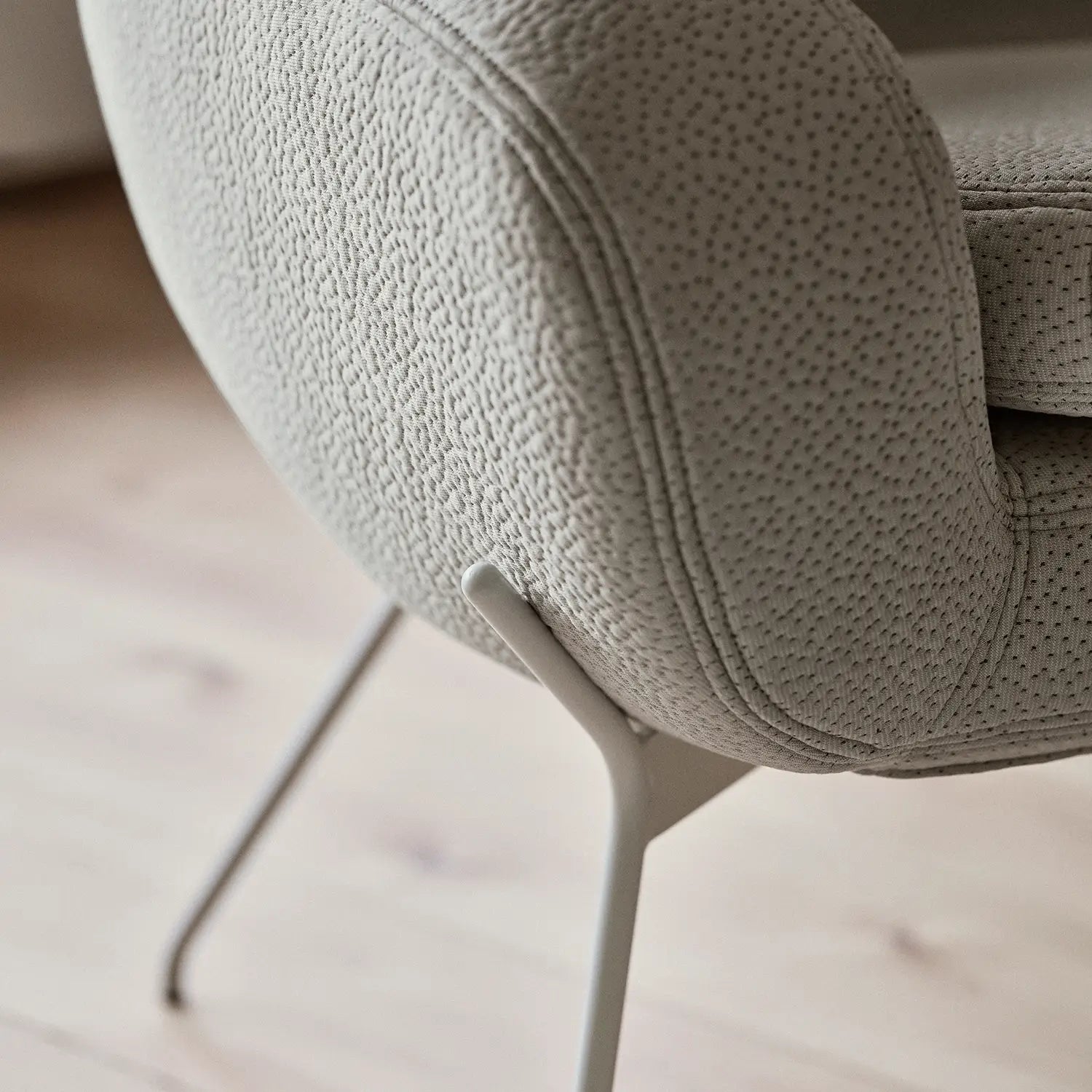 Gros plan sur le Fauteuil NOOMI de Susanne Grønlund, une chaise moderne en tissu texturé beige clair avec des pieds en métal graphique sur un sol en bois clair. Son revêtement doux et pointillé et ses lignes arrondies soulignent son design contemporain.