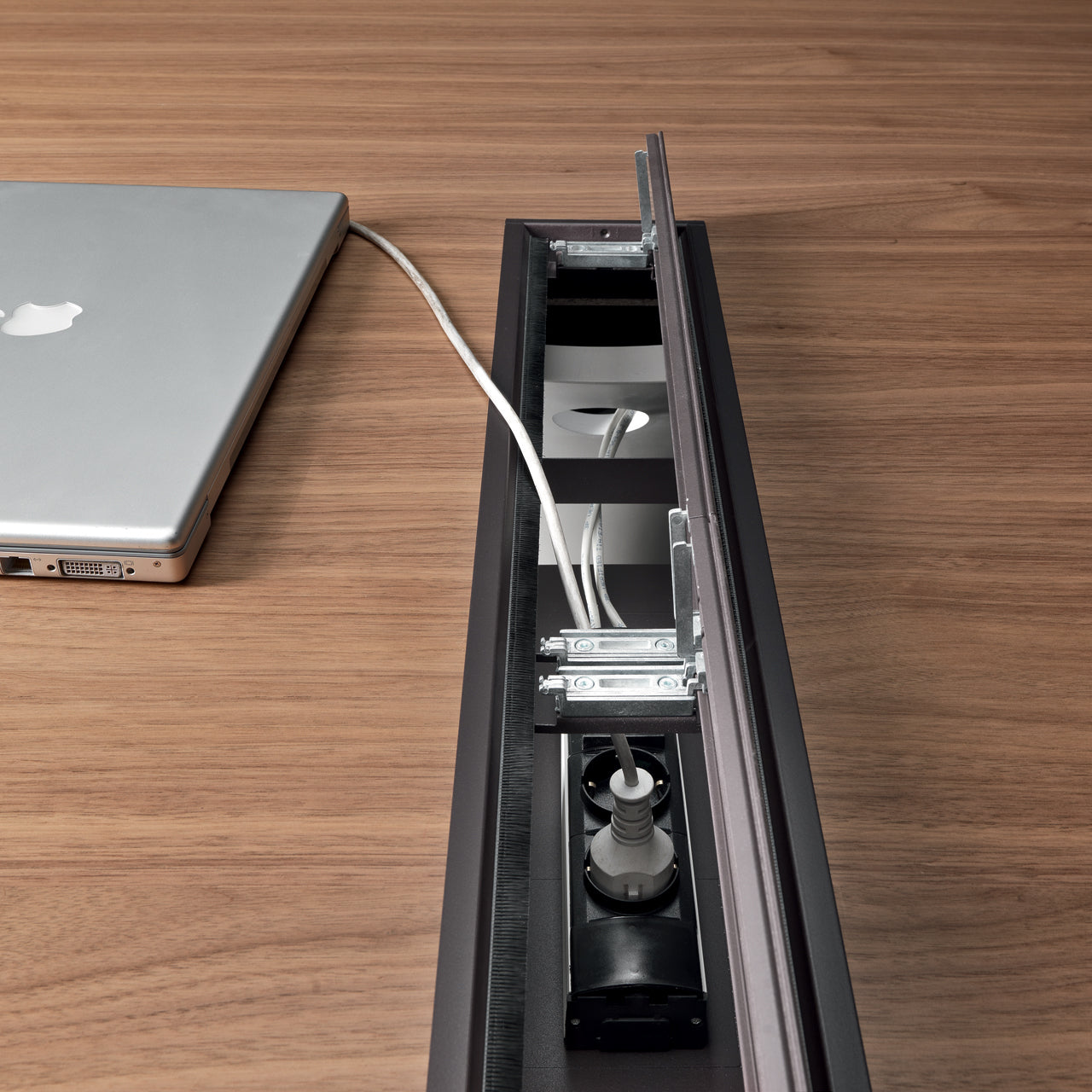 L'Ola d'Attitudes Bureaux est un bureau en bois avec une base elliptique et un couvercle coulissant, partiellement ouvert pour révéler une barre d'alimentation à l'intérieur, où un ordinateur portable argenté est connecté à une prise cachée avec un câble blanc.