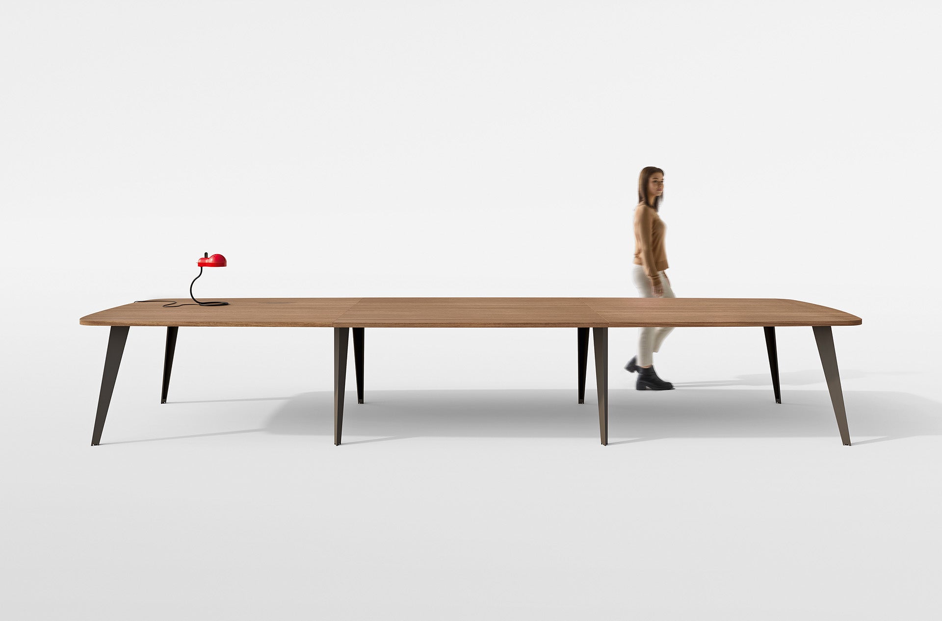 Le Mobilier de direction Pigreco d'Attitudes Bureaux, une longue table en bois avec six pieds noirs, se trouve dans un espace blanc minimal, idéal pour les salles de réunion. Une lampe de bureau rouge est posée à l'une des extrémités, tandis qu'une femme en tenue neutre passe devant.