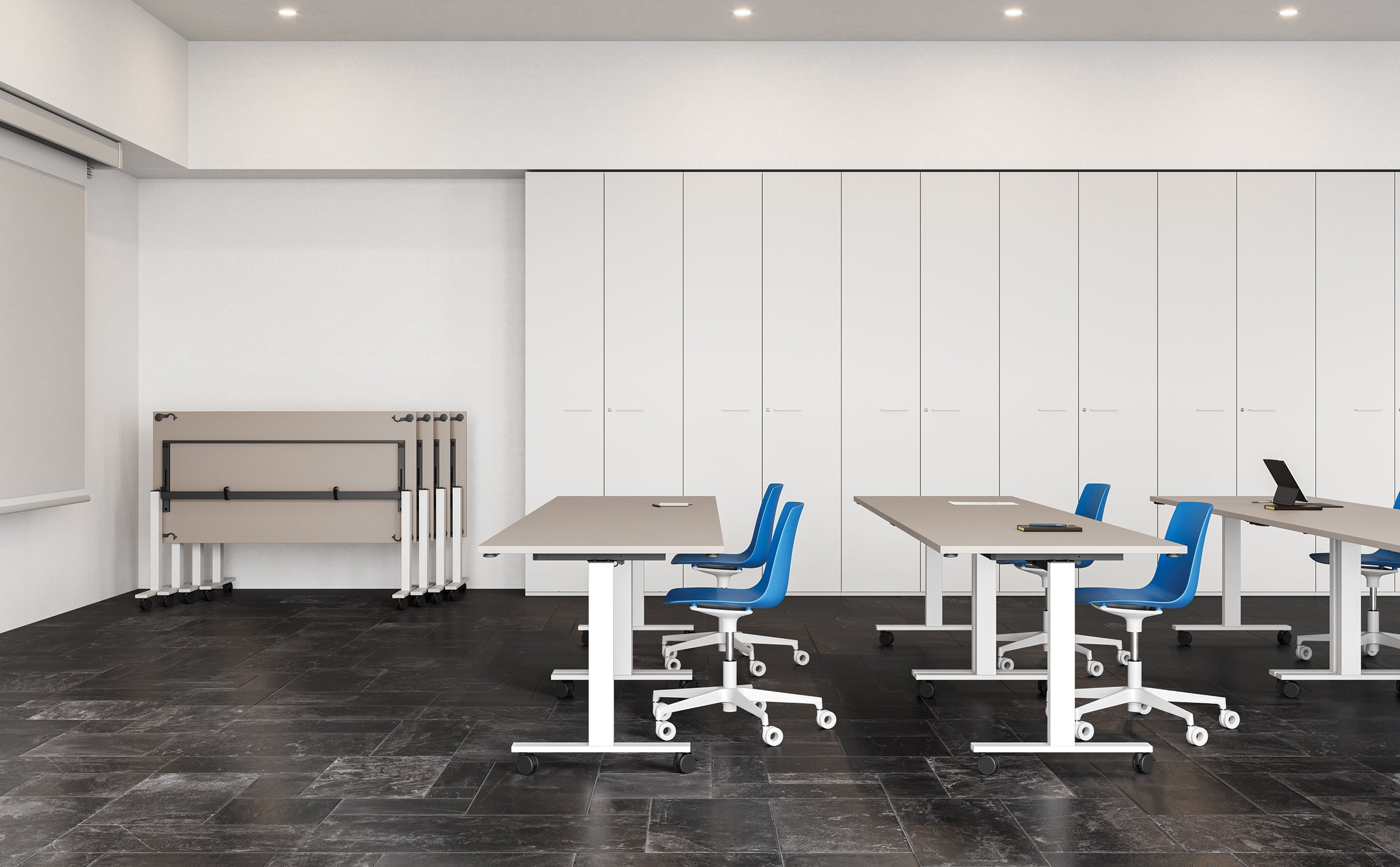 Une salle de classe moderne avec des murs blancs, un sol carrelé noir, des chaises bleues sur roulettes et des tables rectangulaires modulables. Contre le mur du fond se trouvent des chaises empilées et une Attitudes Bureaux Table de réunion pliante sur roulettes Pop Folding. Certaines tables sont équipées d'ordinateurs portables et de cahiers fermés.