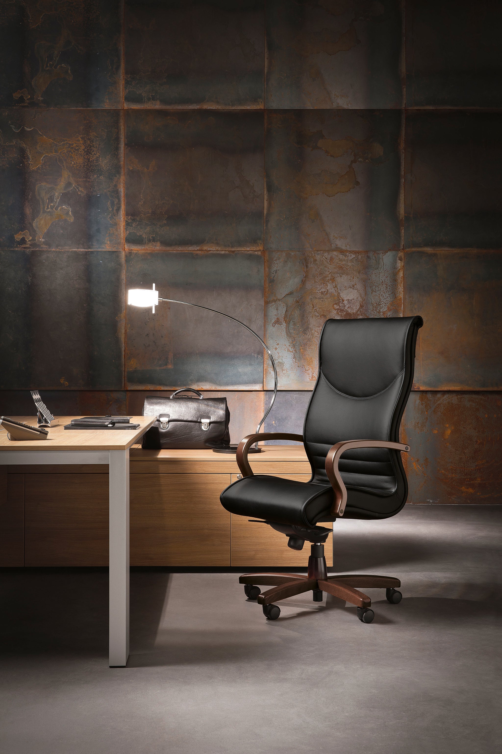 Un bureau moderne avec le Fauteuil de direction Pulchra d'Attitudes Bureaux en cuir noir sur roulettes à côté d'un bureau en bois, avec un porte-documents, un ordinateur portable et une lampe voûtée contre un mur en métal de couleur rouille.