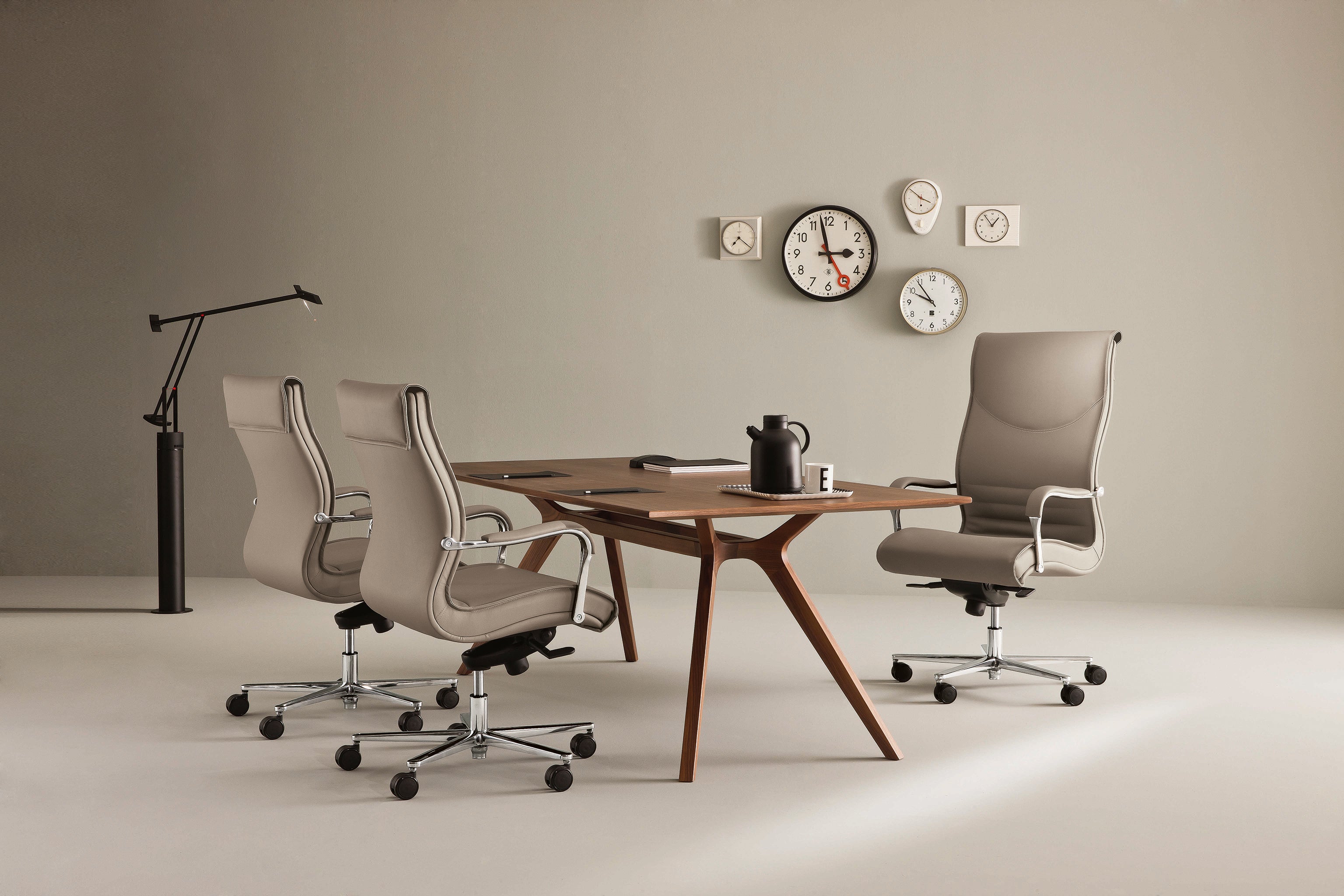Une salle de réunion de bureau moderne avec une table en bois, quatre fauteuils de direction Attitudes Bureaux Pulchra pour un confort ergonomique, une lampe noire, cinq horloges au mur, ainsi qu'une cafetière et un bloc-notes sur la table.