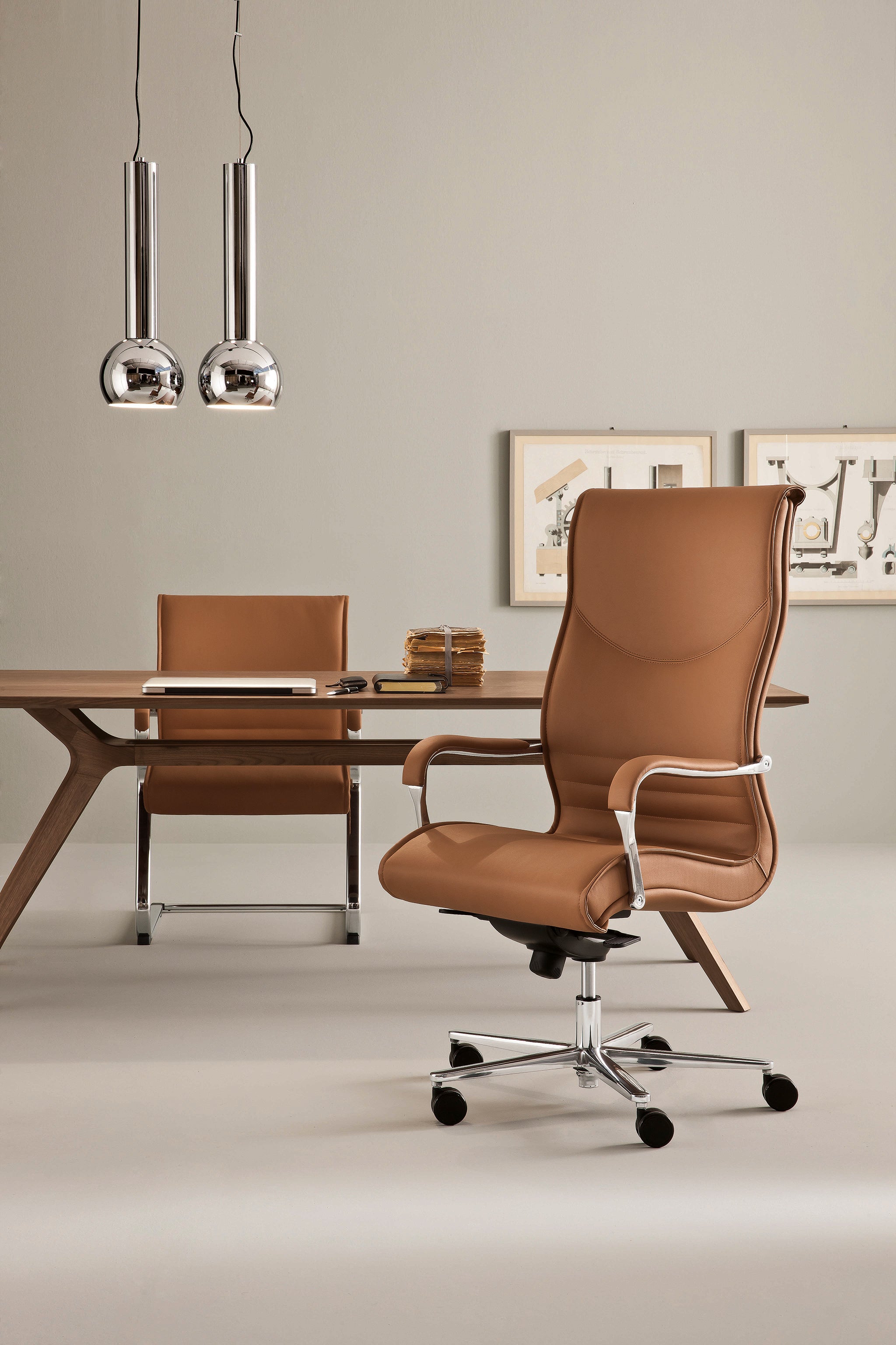 Un bureau moderne équipé du Fauteuil de direction Pulchra d'Attitudes Bureaux en cuir fauve, d'un bureau en bois, de lampes suspendues chromées et d'œuvres d'art encadrées sur des murs beiges - un design élégant, minimaliste et stylé.