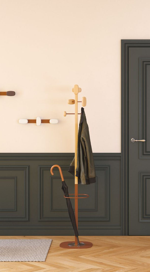 Le Portemanteau SHERWOOD d'Emmanuel Gallina en bois et en acier tient une veste verte et un parapluie noir dans une pièce aux boiseries sombres, aux murs beiges, aux crochets muraux décoratifs et à la porte grise fermée.