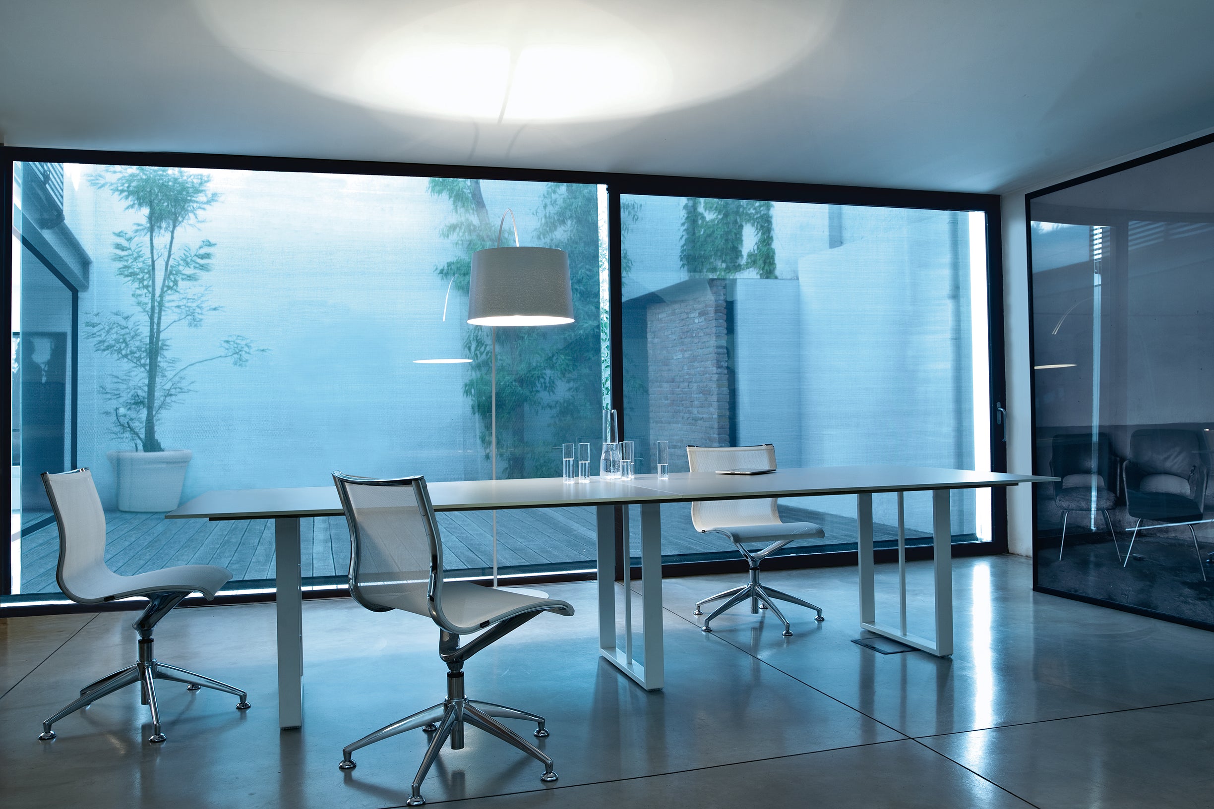 Salle de réunion moderne avec de grandes fenêtres, trois chaises blanches autour d'Attitudes Bureaux Tables de réunion Velvet, de la verrerie sur la table, une lampe suspendue au-dessus, et un patio extérieur avec des plantes visibles à travers les fenêtres.