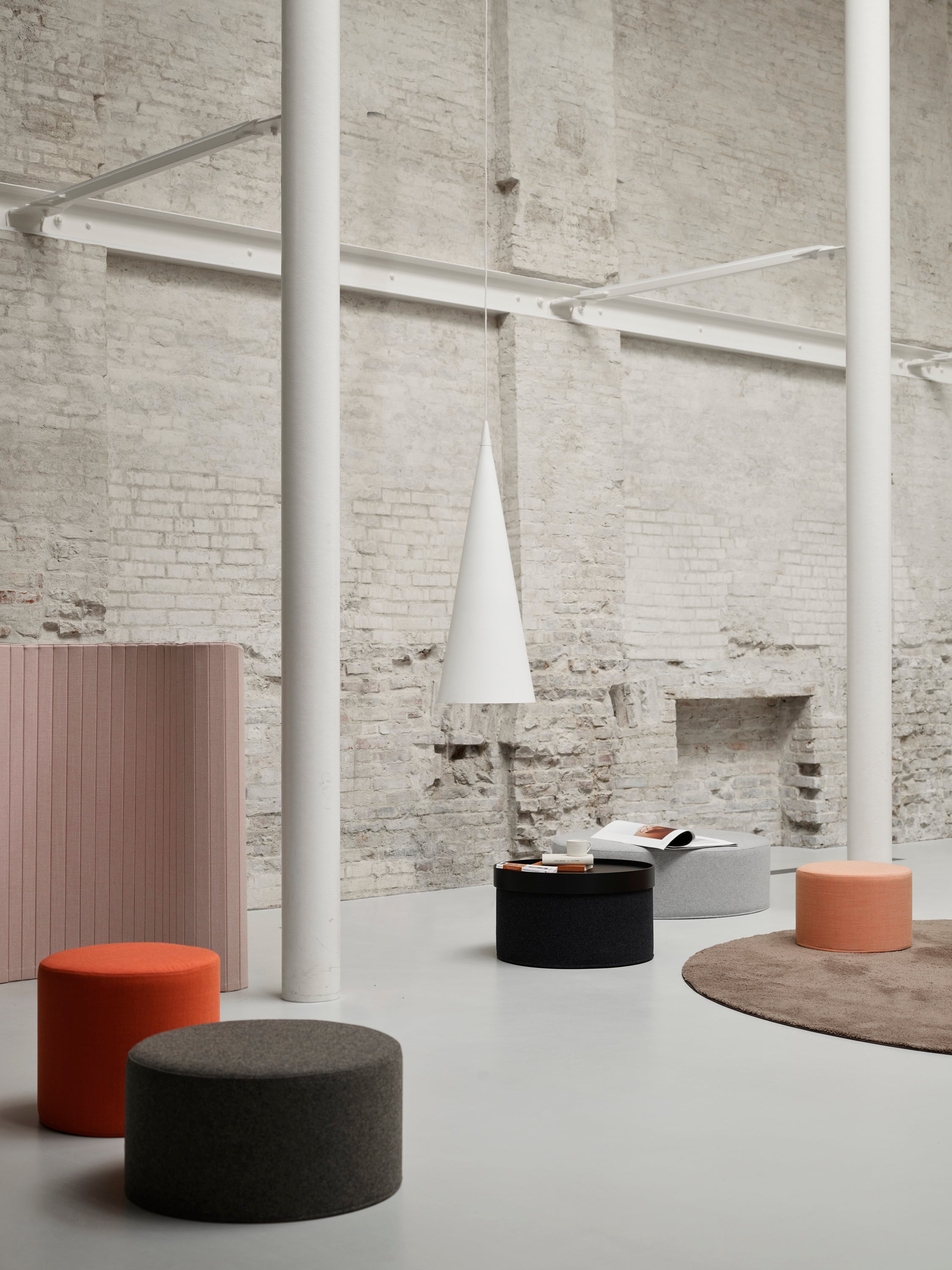 Une pièce minimale et moderne avec des murs en briques apparentes, de hautes colonnes blanches, une table basse, le pouf DRUM multifonction d'Attitudes Bureaux en orange, gris et noir, un tapis rond marron, une lampe suspendue à cône blanc et un livre ouvert sur une table noire.