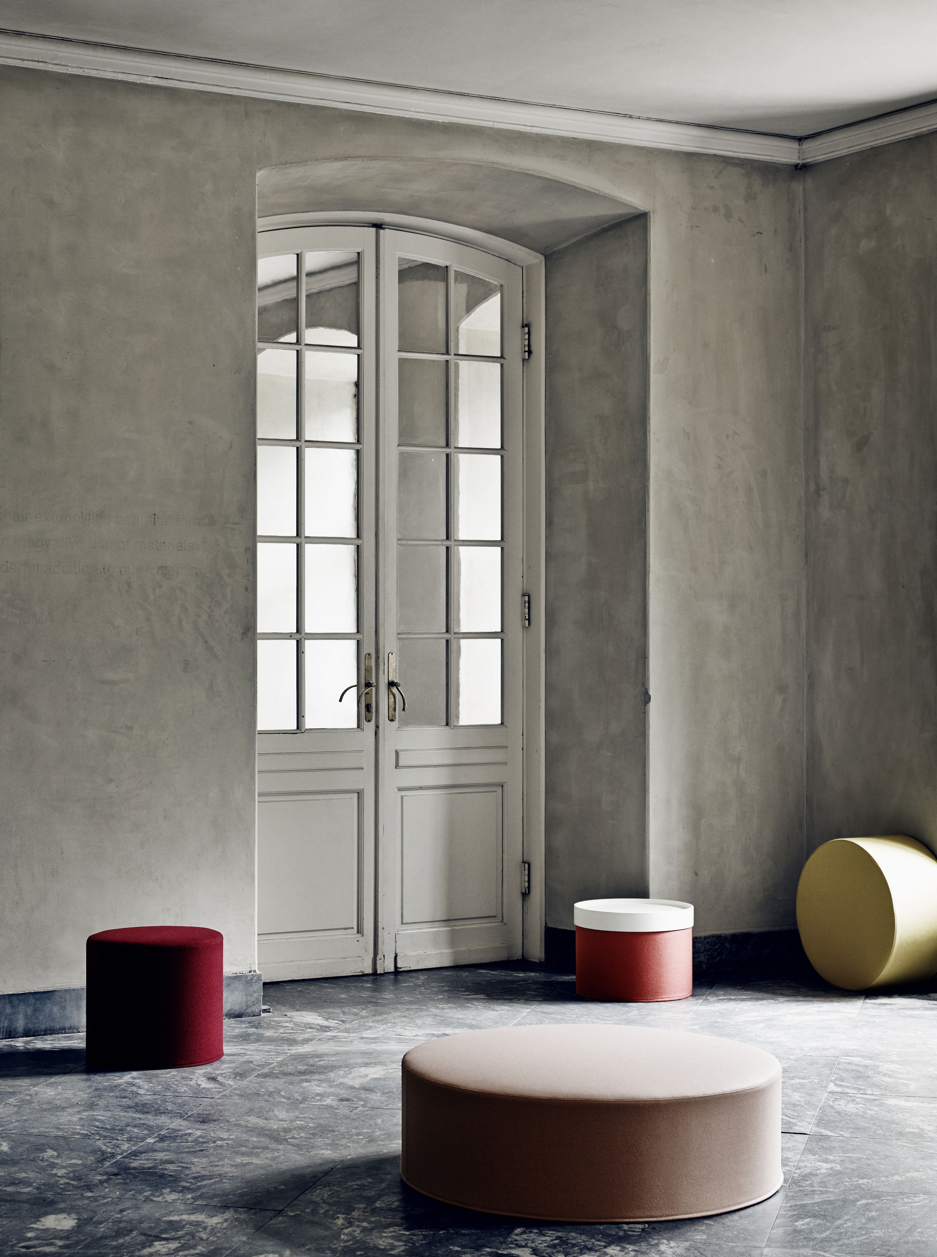 Pièce minimaliste avec murs et sol gris, hautes doubles portes vitrées, et trois poufs DRUM multifonction d'Attitudes Bureaux, utilisés comme ottomans ou table basse, mettant en valeur l'élégant mobilier modulable dans l'ensemble de l'espace.