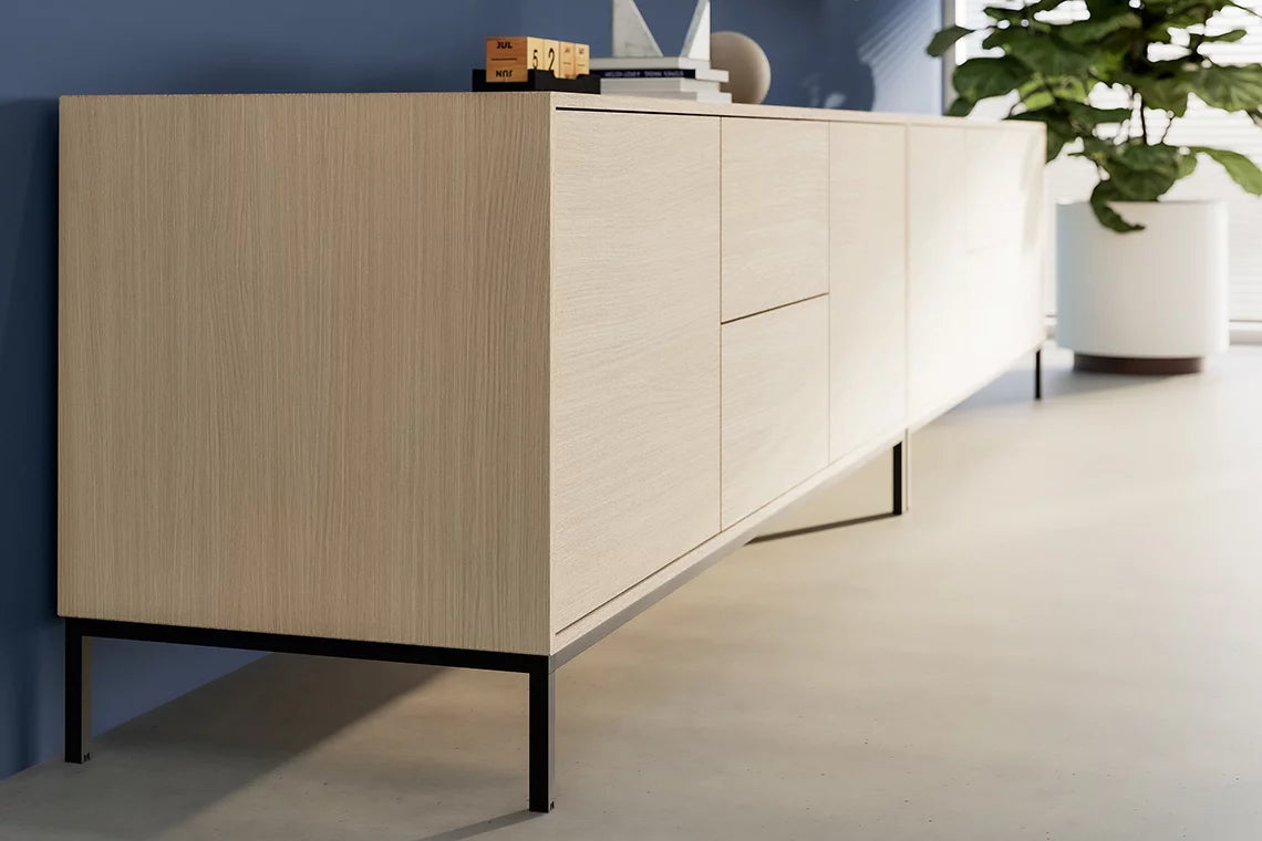 Le Buffet Quadra d'Attitudes Bureaux, un buffet moderne en bois clair avec des pieds en métal noir, se dresse contre un mur bleu. La lumière du soleil à travers les stores met en valeur le design épuré de ce rangement pour bureaux, complété par des éléments de décoration et une grande plante verte.