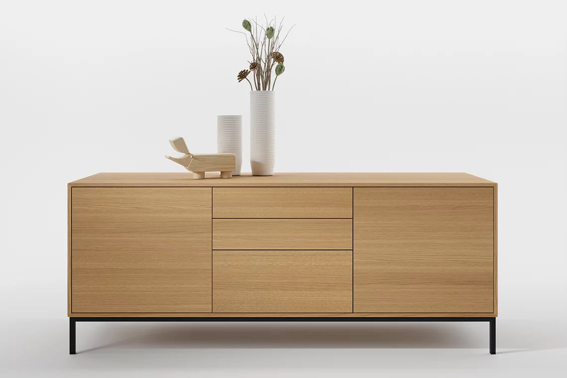 Le Buffet Quadra d'Attitudes Bureaux est un buffet de bureau moderne en bois aux lignes épurées, composé de deux armoires, trois tiroirs sur un socle en métal noir, surmonté de deux vases blancs contenant des plantes séchées et d'un petit objet décoratif en bois - parfait pour le rangement de bureau.