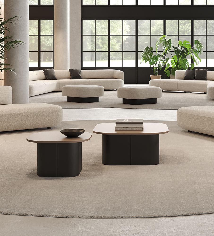 Un salon moderne et spacieux avec de grandes fenêtres, des canapés incurvés beige clair, des tables basses Ramos Bassols Afina, des tables basses noires et beiges, un tapis rond clair et des plantes en pot vertes. La lumière naturelle remplit la pièce.