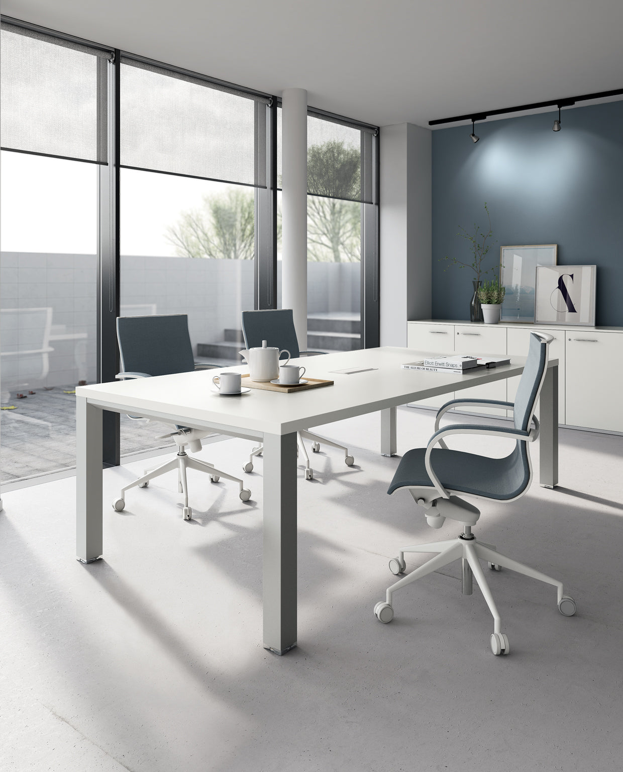 Bureau moderne avec de grandes fenêtres, une table Attitudes Bureaux Tables de réunion Asset, trois chaises bleues à roulettes, deux tasses à café, un carnet de notes et un décor minimaliste. La lumière naturelle remplit la pièce avec les arbres visibles à l'extérieur.