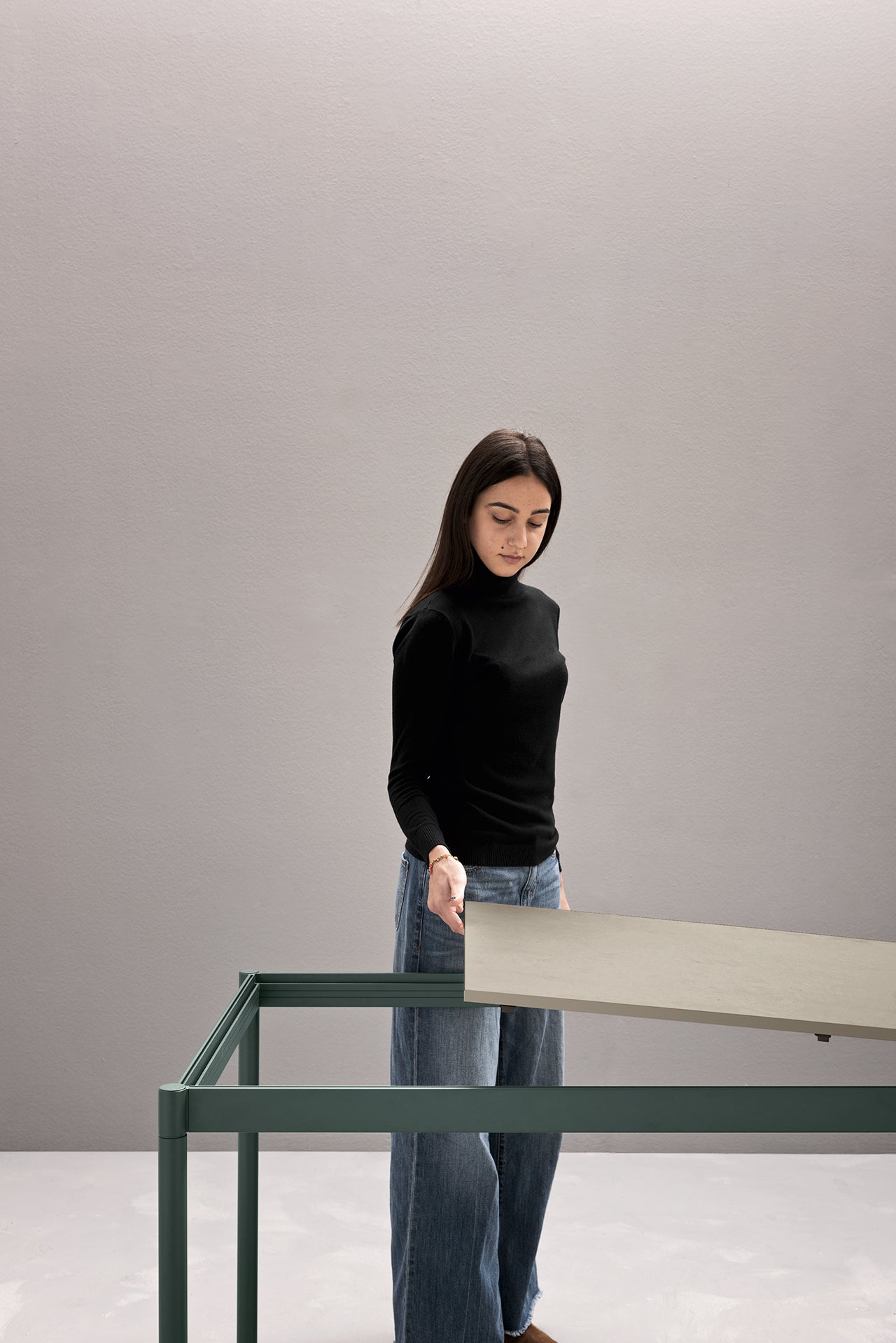 Une femme aux longs cheveux noirs, vêtue d'un haut noir et d'un jean bleu, fait la démonstration de Fluido, mobilier modulaire transformable d'Attitudes Bureaux en plaçant un panneau lumineux sur une table à cadre métallique vert sur un fond gris uni.