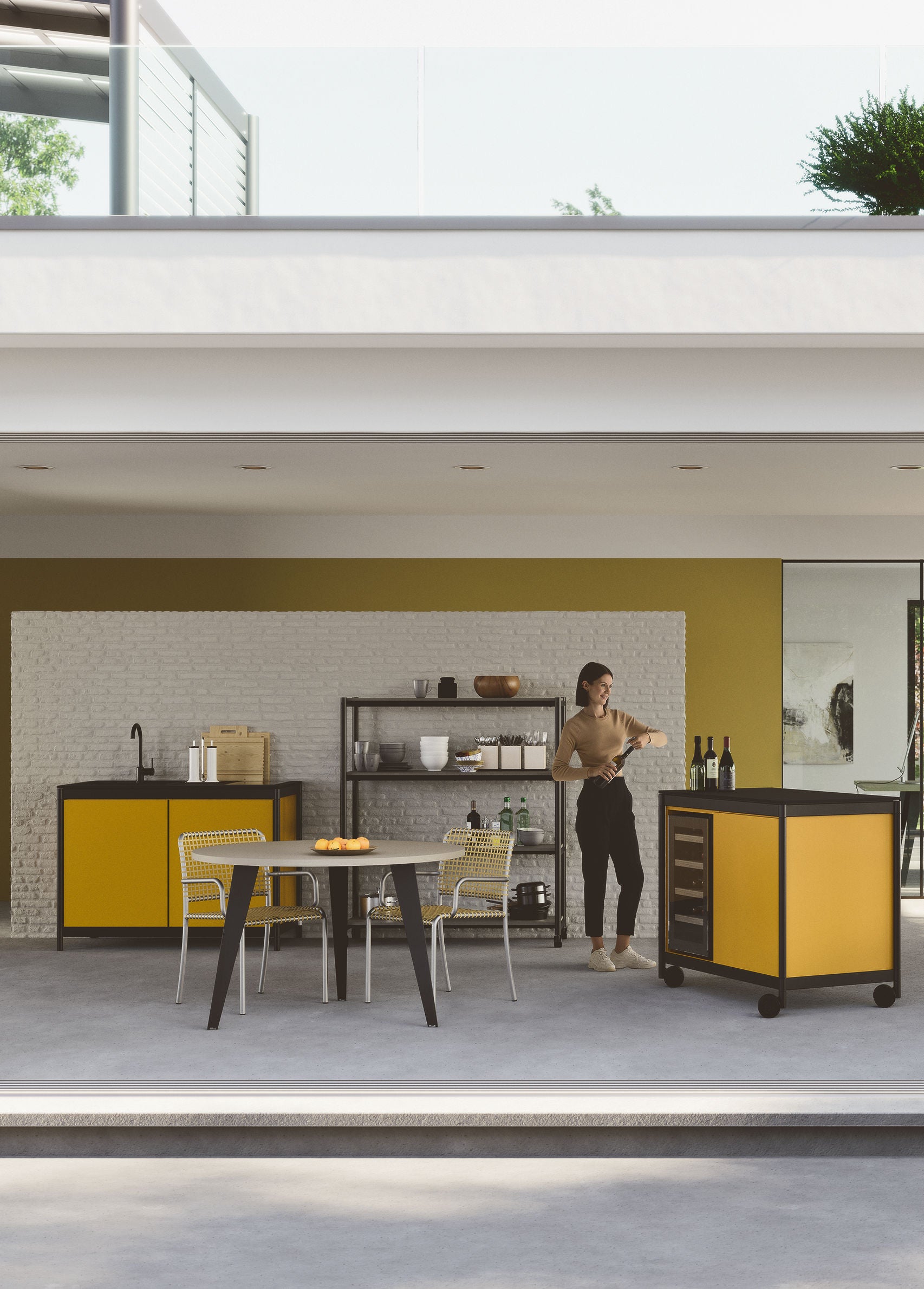 Une femme se tient dans une cuisine moderne intérieure-extérieure avec des armoires jaunes, une petite table à manger et la Kitchenette Fluido® d'Attitudes Bureaux, avec des étagères contenant des articles de cuisine sur un mur de briques blanches et de jaune moutarde.