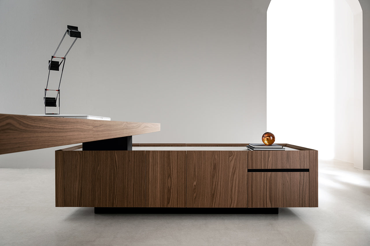Bureau minimaliste avec le bureau moderne en bois Kyo d'Attitudes Bureaux, défini par des lignes épurées. Il comprend une lampe noire, des livres empilés et une sphère en verre ambré. Des matériaux de qualité supérieure et une porte cintrée lumineuse complètent le look.