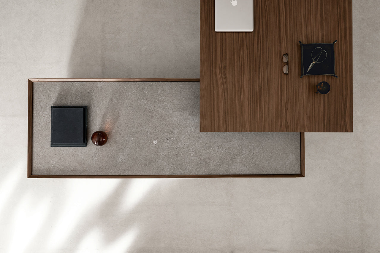 Espace de travail minimaliste vu du dessus, comprenant le bureau Kyo d'Attitudes Bureaux, avec un ordinateur portable, des lunettes et un plateau, à côté d'une surface en béton avec un cahier fermé et une sphère marron, le tout sur un sol gris clair.