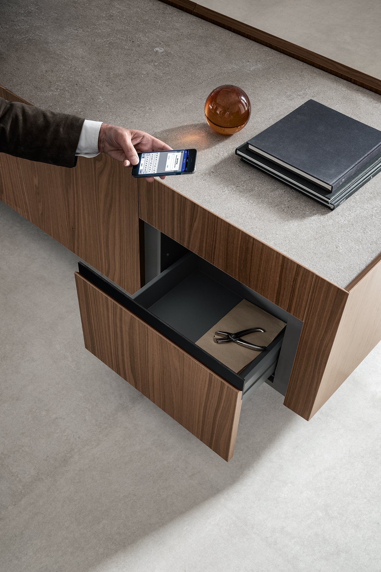 Une personne munie d'un smartphone se tient près du bureau flottant en bois Kyo d'Attitudes Bureaux, fabriqué à partir de matériaux de première qualité, qui comporte un tiroir ouvert avec des lunettes et des livres, ainsi qu'une sphère en verre sur son comptoir en béton.